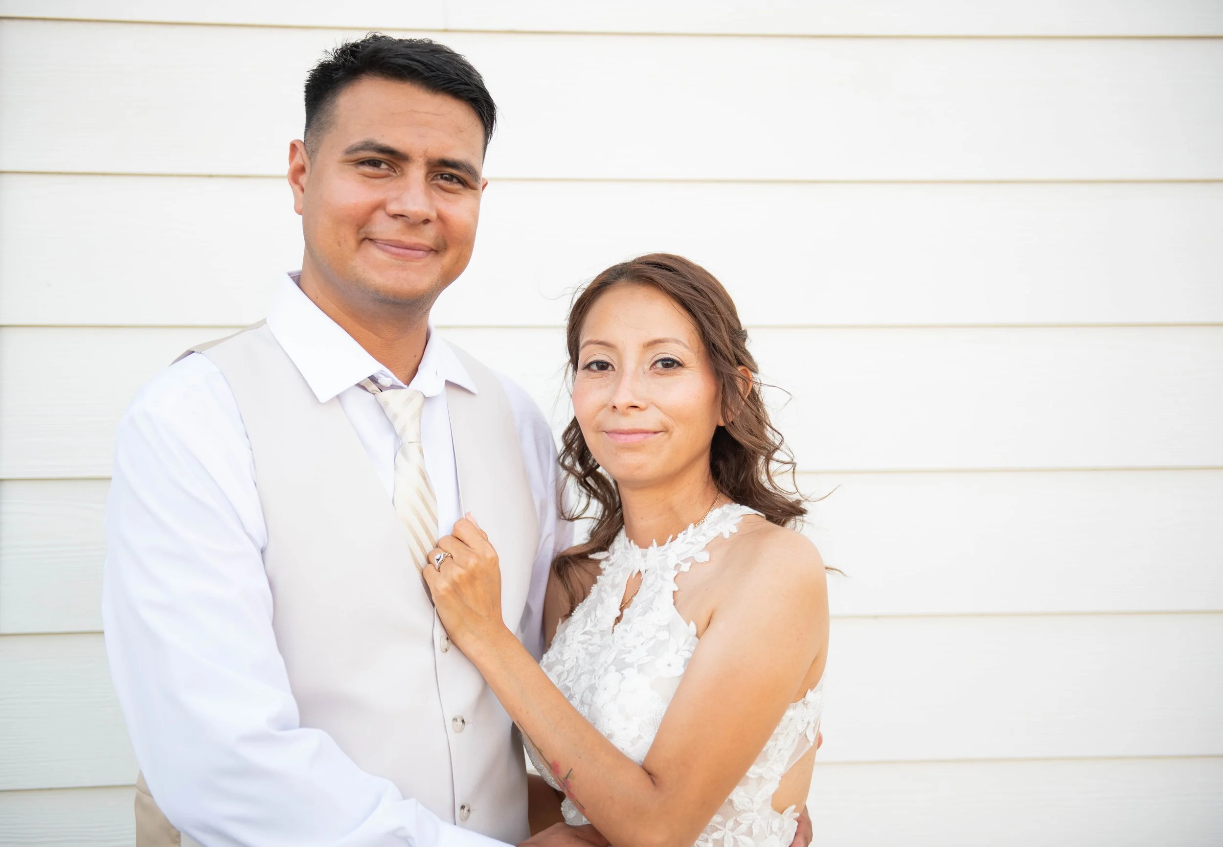 A young man and woman, both dressed in white, standing close together outdoors against a white horizontal siding wall. The woman is wearing a white lace dress and has her hand on the man's chest, showing her wedding ring. They are smiling softly.