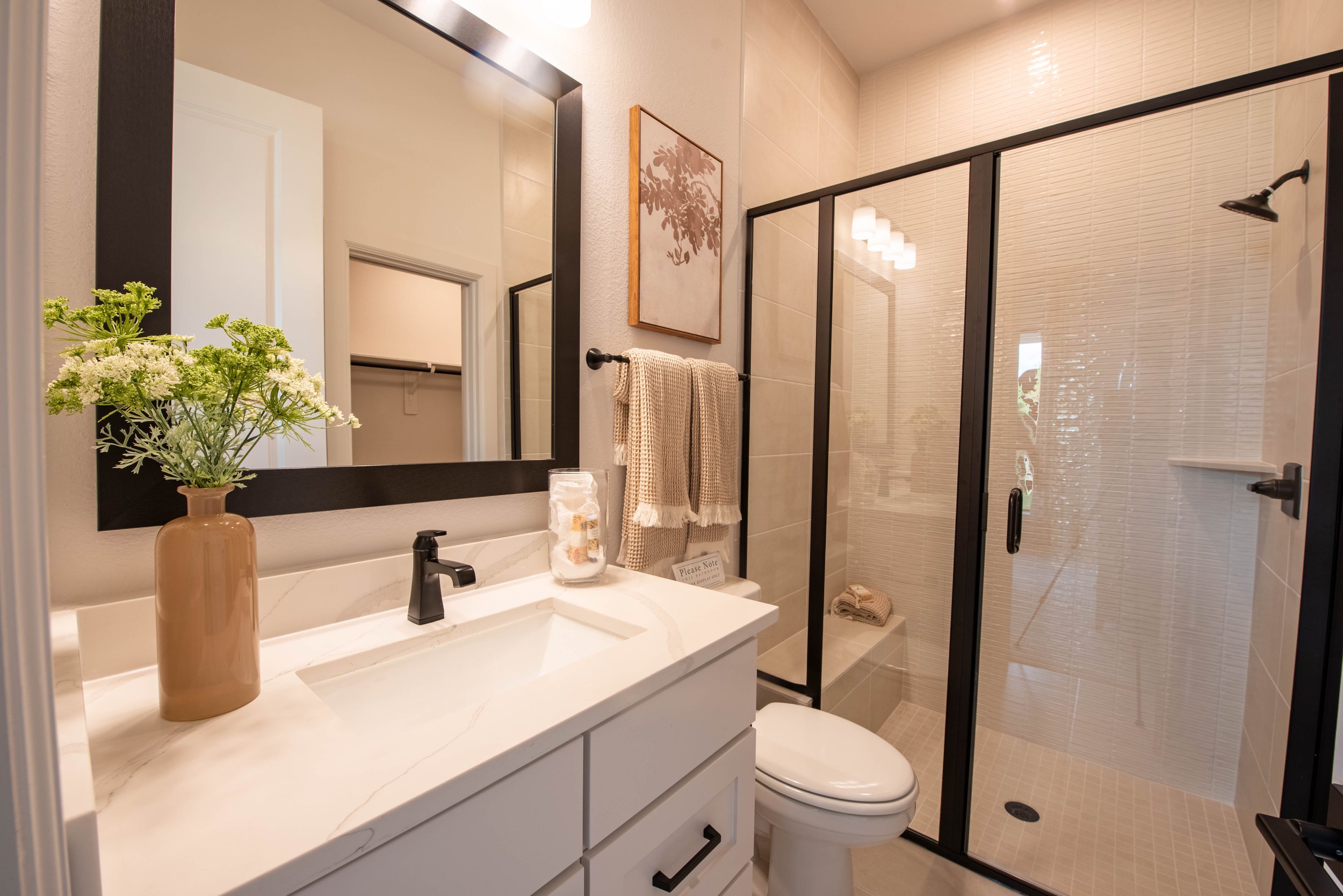 Modern bathroom with a white vanity, black faucet, mirror, framed artwork, towel rack with beige towel, glass shower enclosure, and a toilet.
