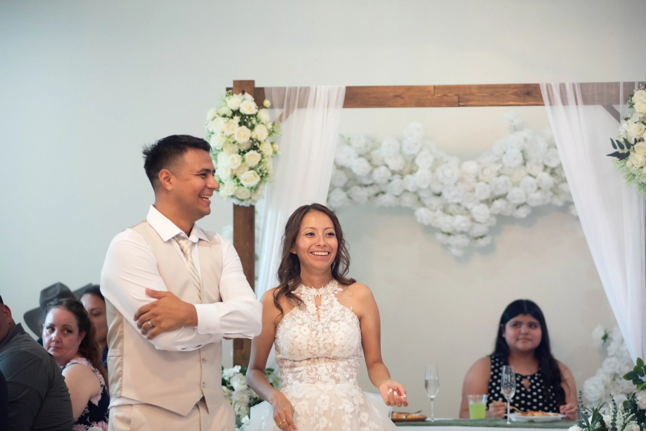 A couple standing at a wedding ceremony, with the groom smiling and the bride in a white lace wedding dress inside a decorated ceremony space with floral arrangements and guests seated around.