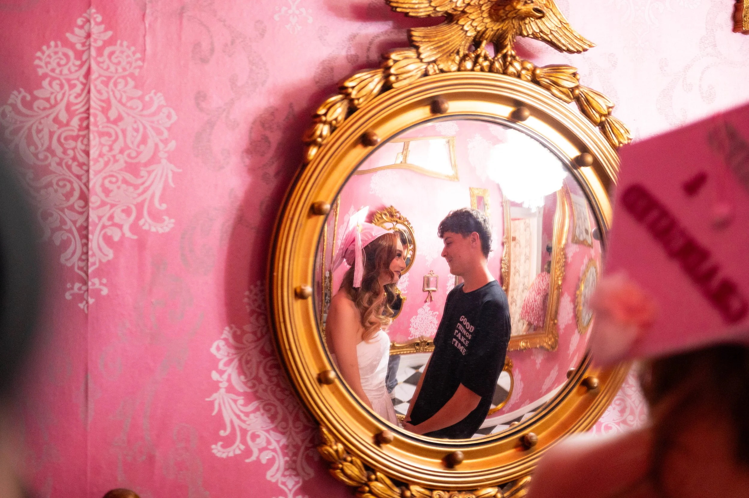 A couple is seen smiling and looking at each other in a mirror framed with gold, surrounded by pink wallpaper with white floral patterns. The woman has long curly hair and is wearing a strapless white top, while the man has short dark hair and is wea