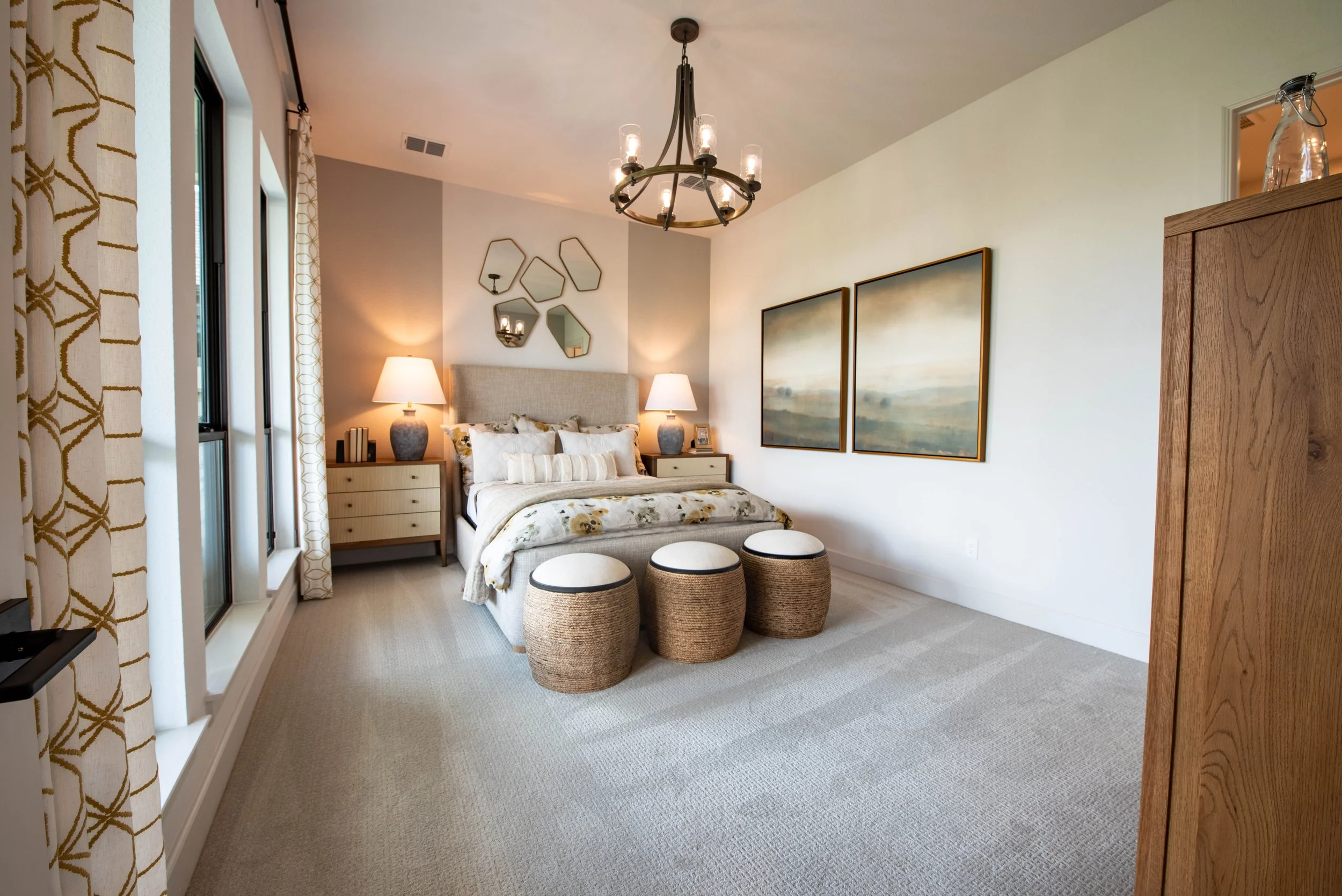 A bedroom with a bed, two nightstands with lamps, three woven stools, wall art, and a chandelier.