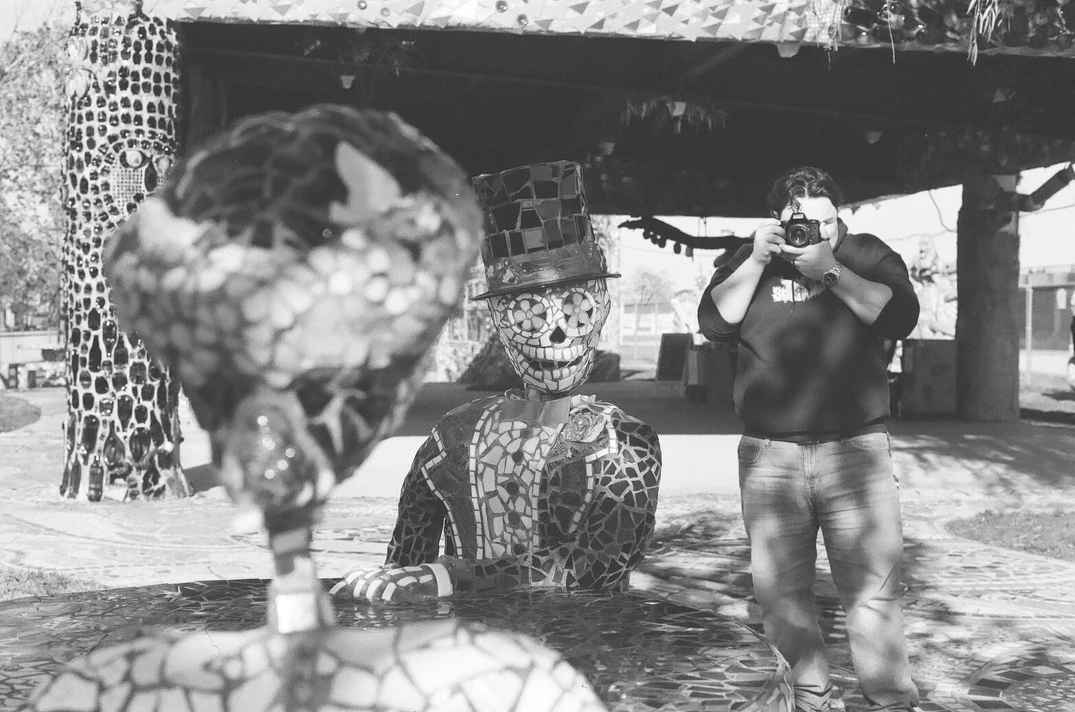 Person taking a photograph of a mosaic sculpture of a skeleton with a top hat outdoors.