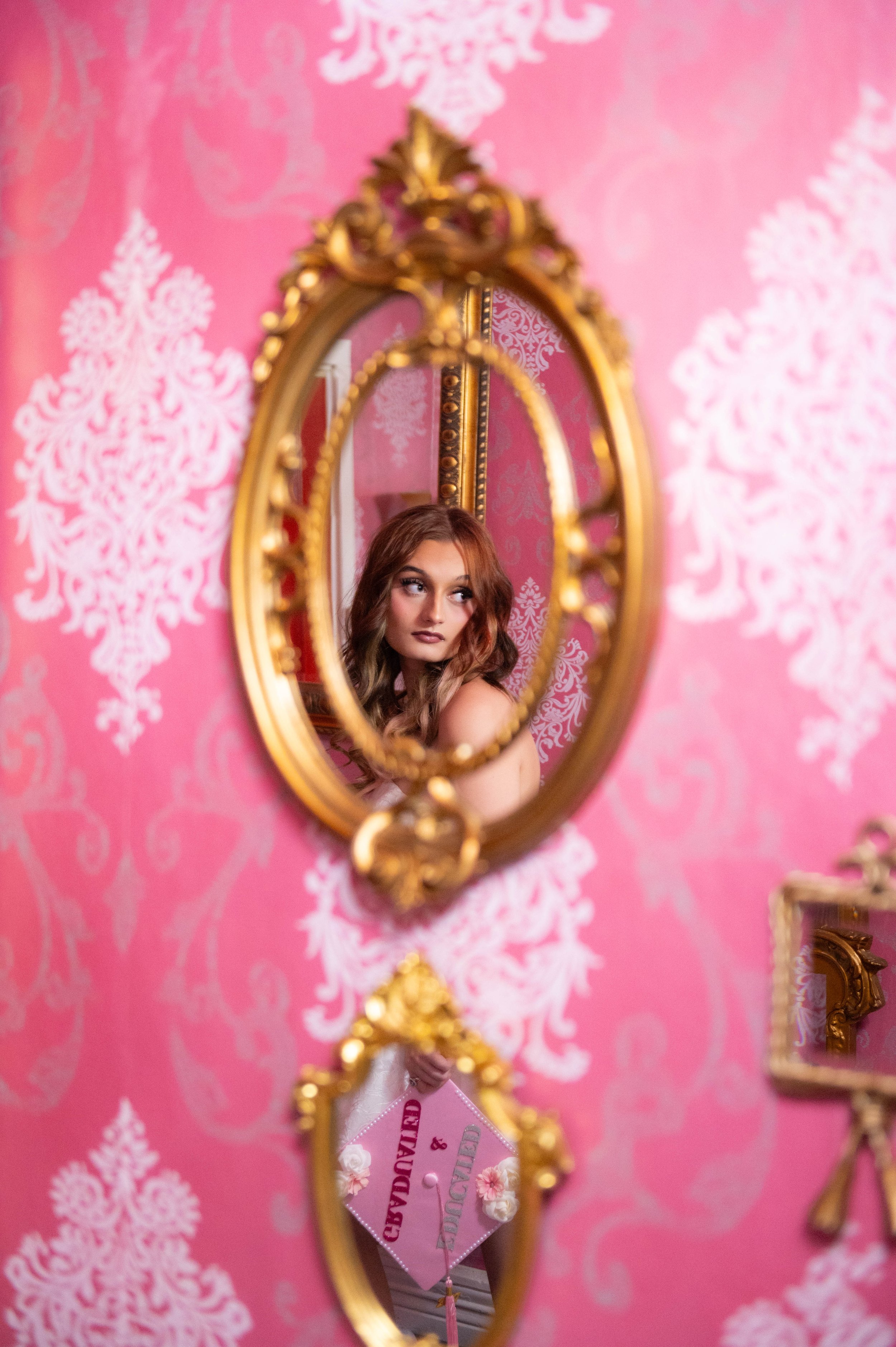 A young woman with red hair looking at her reflection in an ornate gold-framed oval mirror, mounted on a pink wallpapered wall with white floral patterns. The woman appears to be in a dressing room, and there's a pink ticket with white flowers visibl