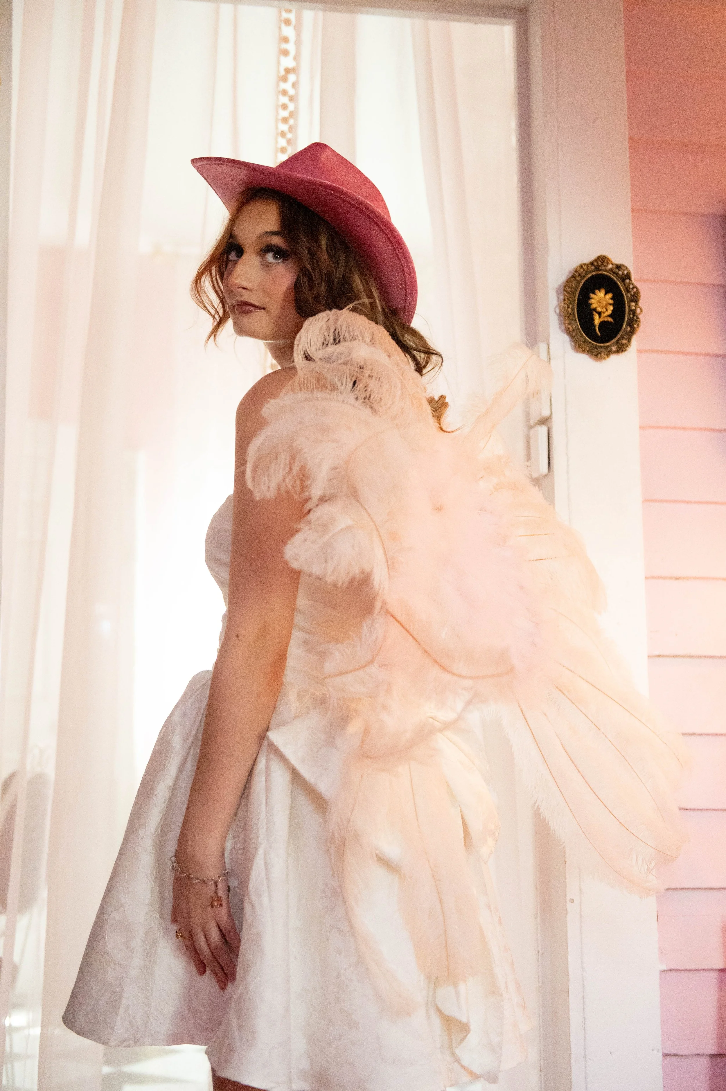 A woman dressed in a white, strapless gown with pink feathered angel wings and a pink wide-brimmed hat, standing indoors near a doorway with pink walls and light curtains.
