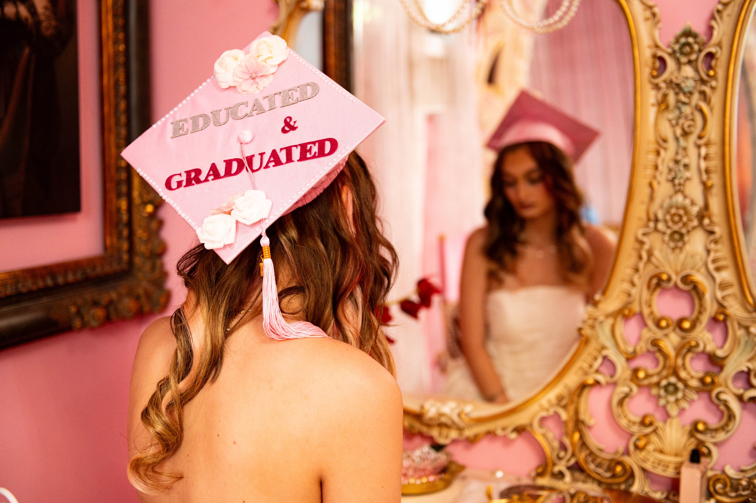 Graduate woman wearing pink cap with 'Educated & Graduated' written on it, standing in front of ornate mirror and pink wall.