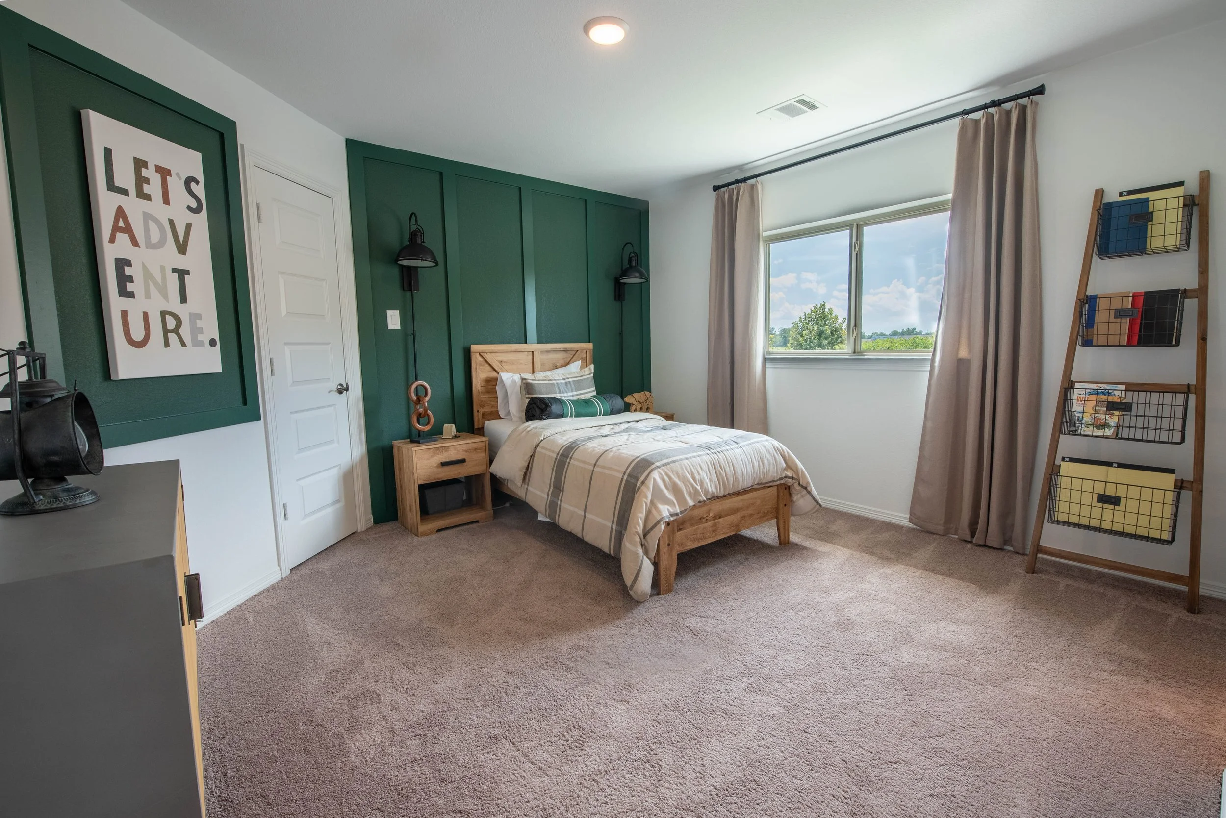 A bedroom with a wooden bed, green accent wall, large window with beige curtains, and modern decor including a framed poster reading 'Let's Adventur' and a wooden ladder shelf with wire baskets.