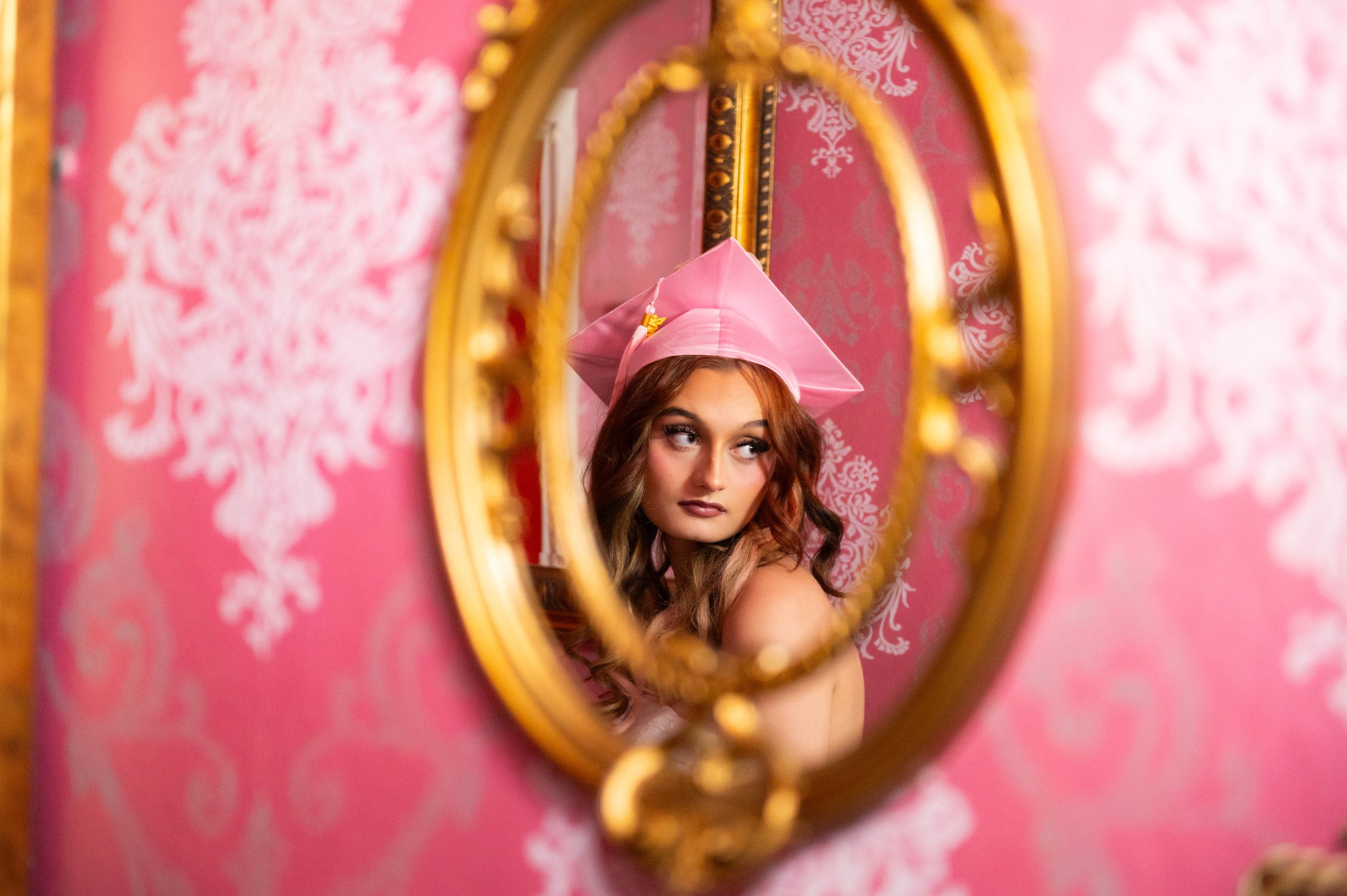 Young woman wearing a pink graduation cap, seen through an ornate mirror with a pink patterned wall in the background.