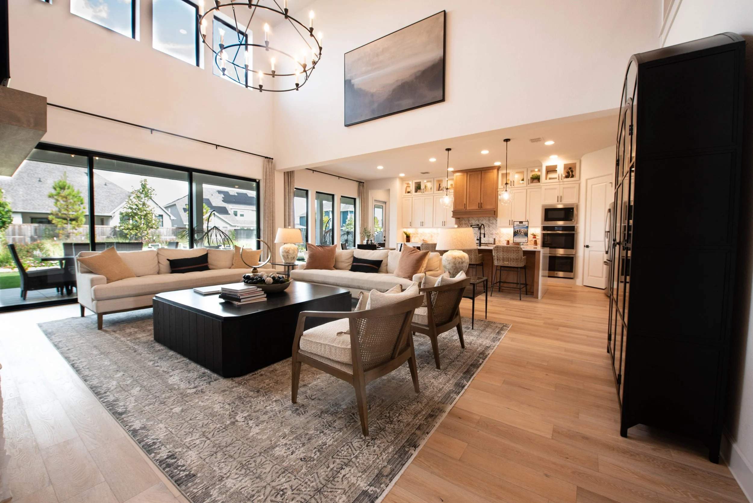 Open-concept living room and kitchen with large glass windows, white sofas, a black coffee table, rattan chairs, and a modern chandelier in a bright, contemporary home.