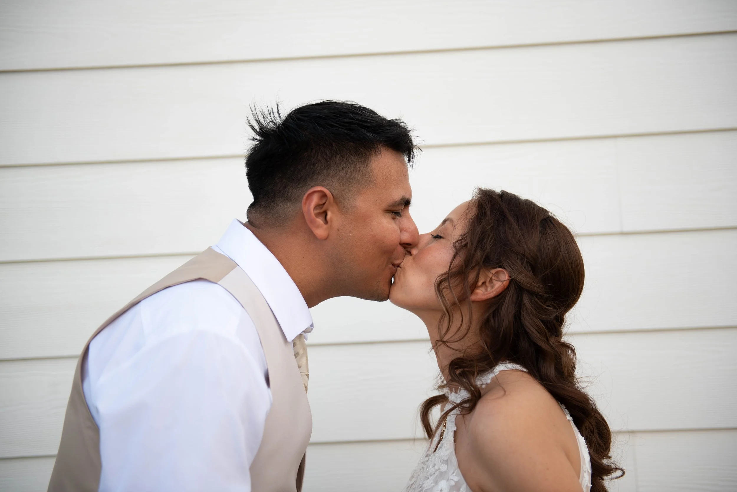 A man and woman sharing a kiss in front of a white wooden wall, with the man wearing a suit and the woman in a lace dress.