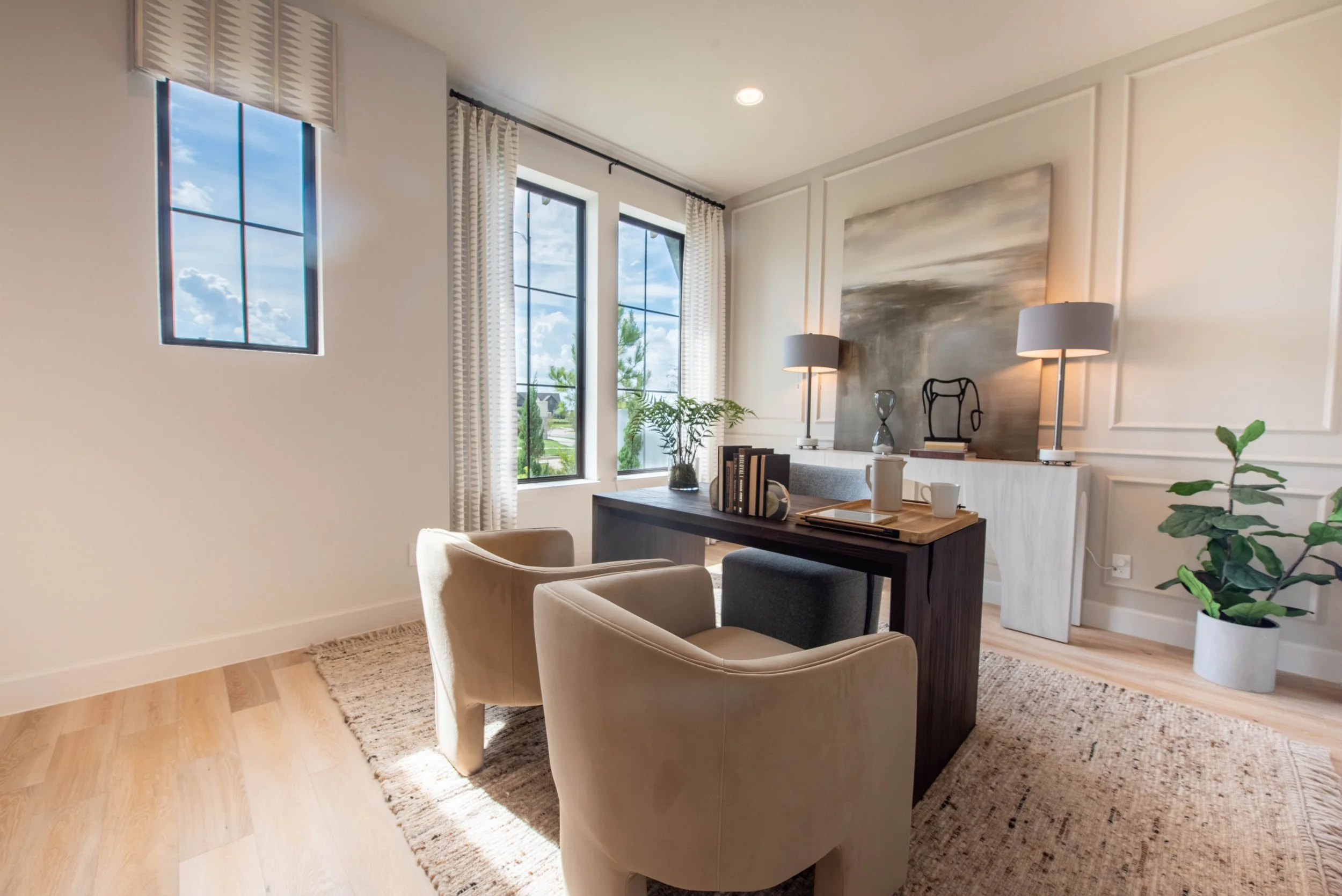 A cozy, modern living room with large windows letting in natural light, white curtains, a wooden table with books and a tray, cream armchairs, green plants, and a textured beige rug.
