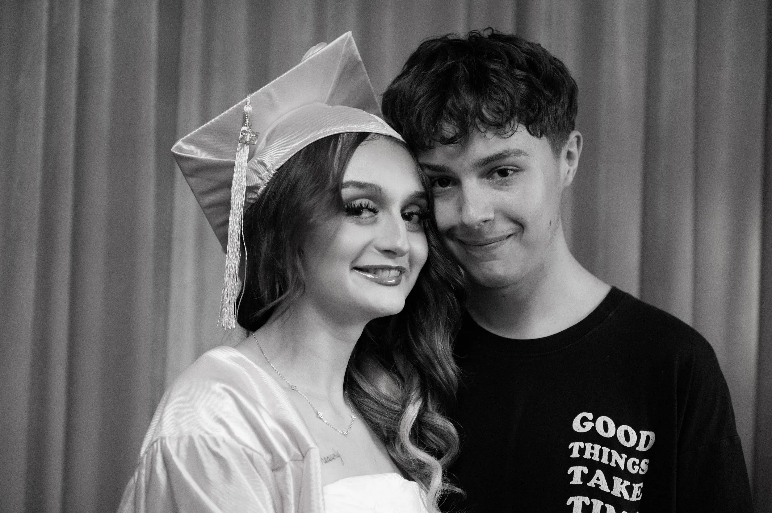 A young woman in a cap and gown, and a young man in a T-shirt, both smiling and holding close in a graduation celebration.