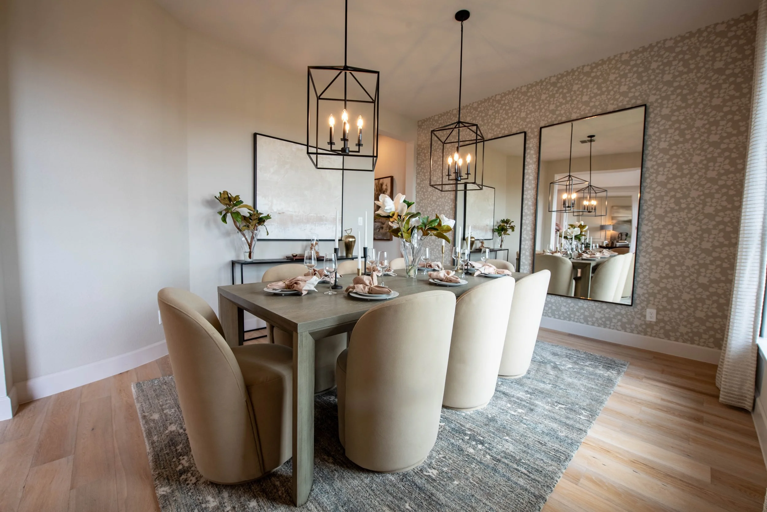 A dining room with a wooden table set for a meal, surrounded by cream-colored upholstered chairs. The table has candles, glassware, and napkins. There are large mirrors on the wall reflecting the room, and modern black pendant lights hanging from the