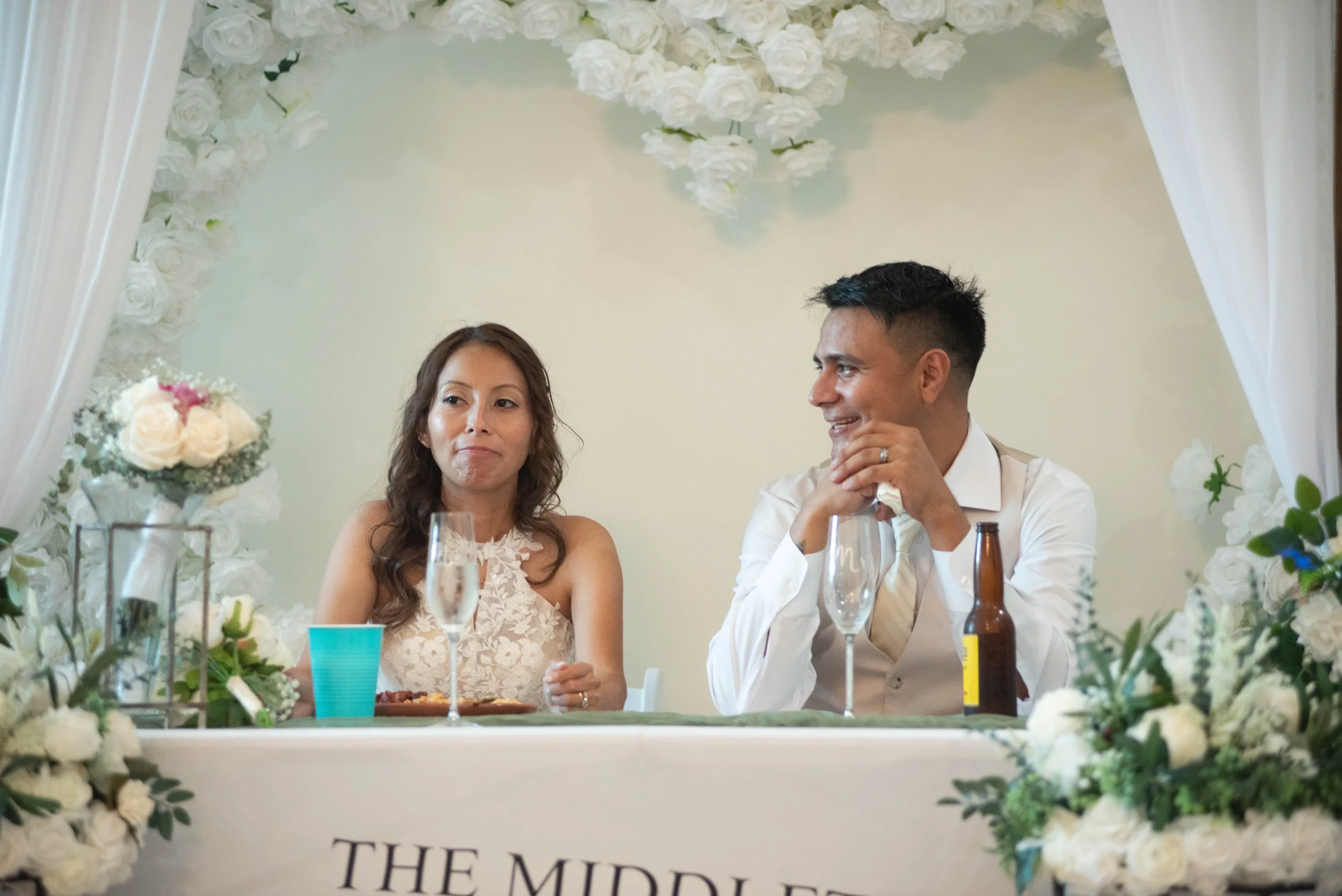 A bride and groom sit at a decorated reception table, surrounded by white flowers and greenery, during their wedding celebration.