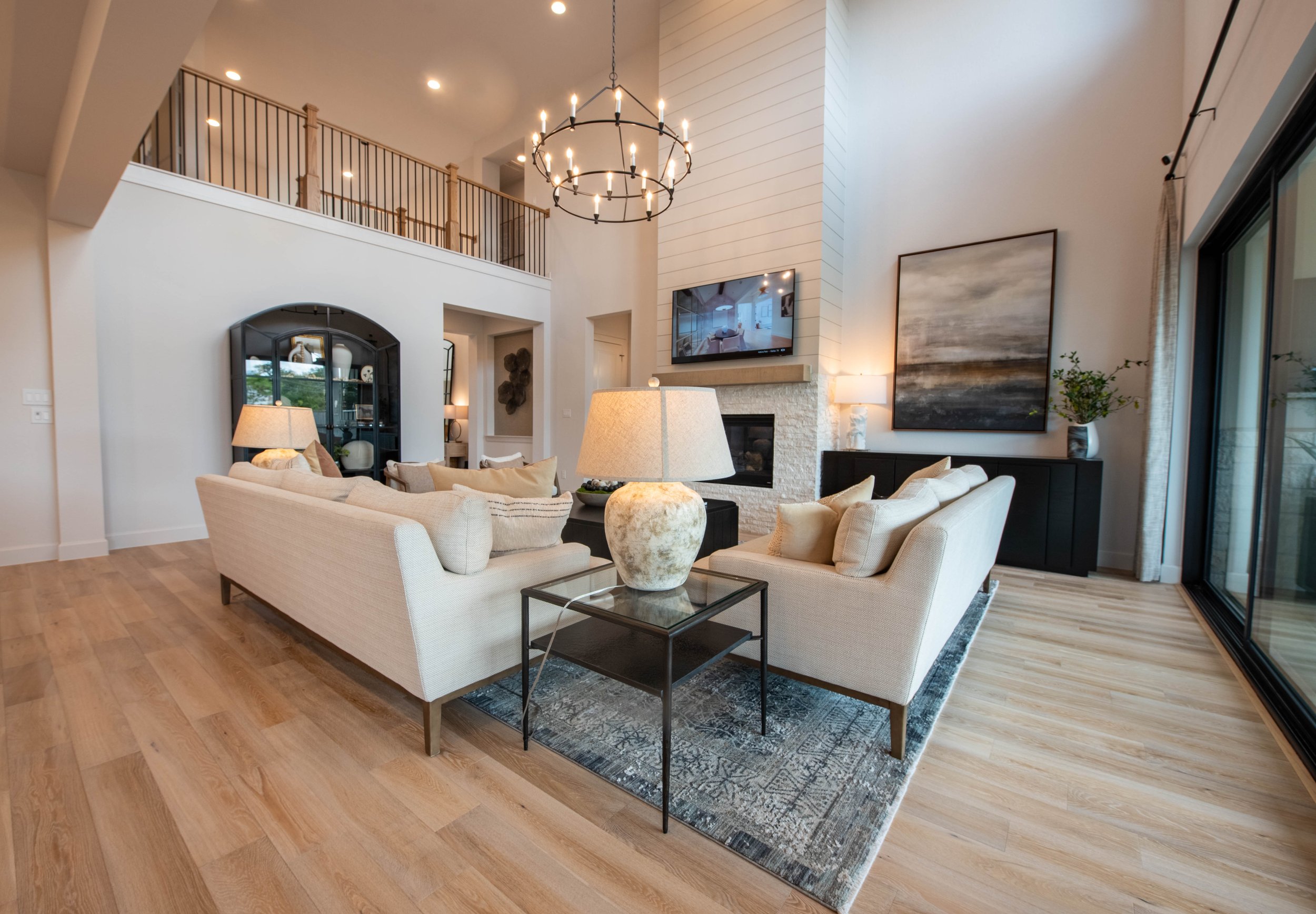 Living room with white sofas, a glass-topped side table with a large stone base, a decorative rug, a flat-screen TV mounted above a fireplace, large window with curtains, contemporary artwork on the wall, and a chandelier hanging from the high ceilin