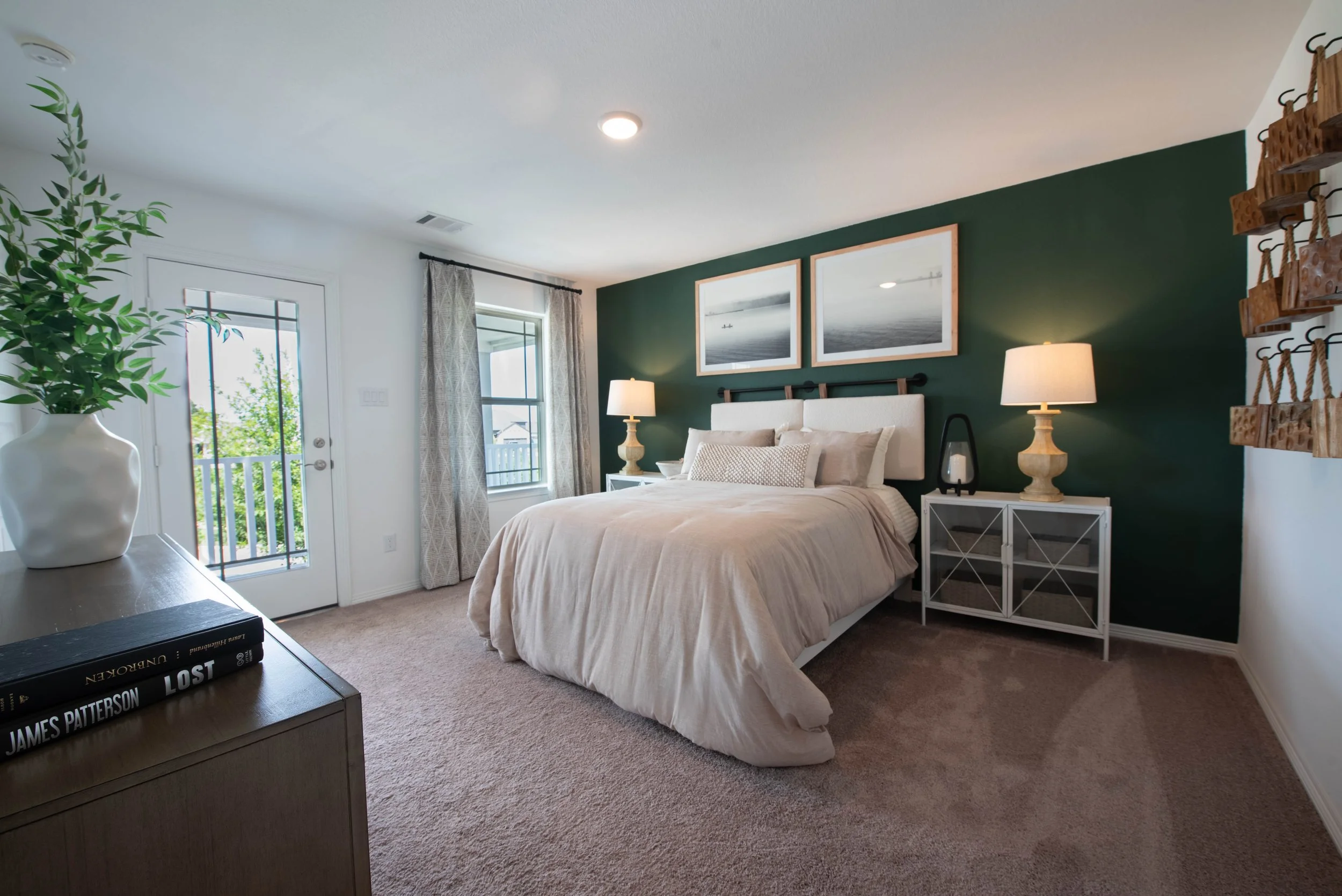 Bedroom with green accent wall, large bed with beige bedding, two nightstands with lamps, framed black-and-white photos above the bed, window with curtains, door with glass panel, decorated shelves, and a large potted plant in the corner.