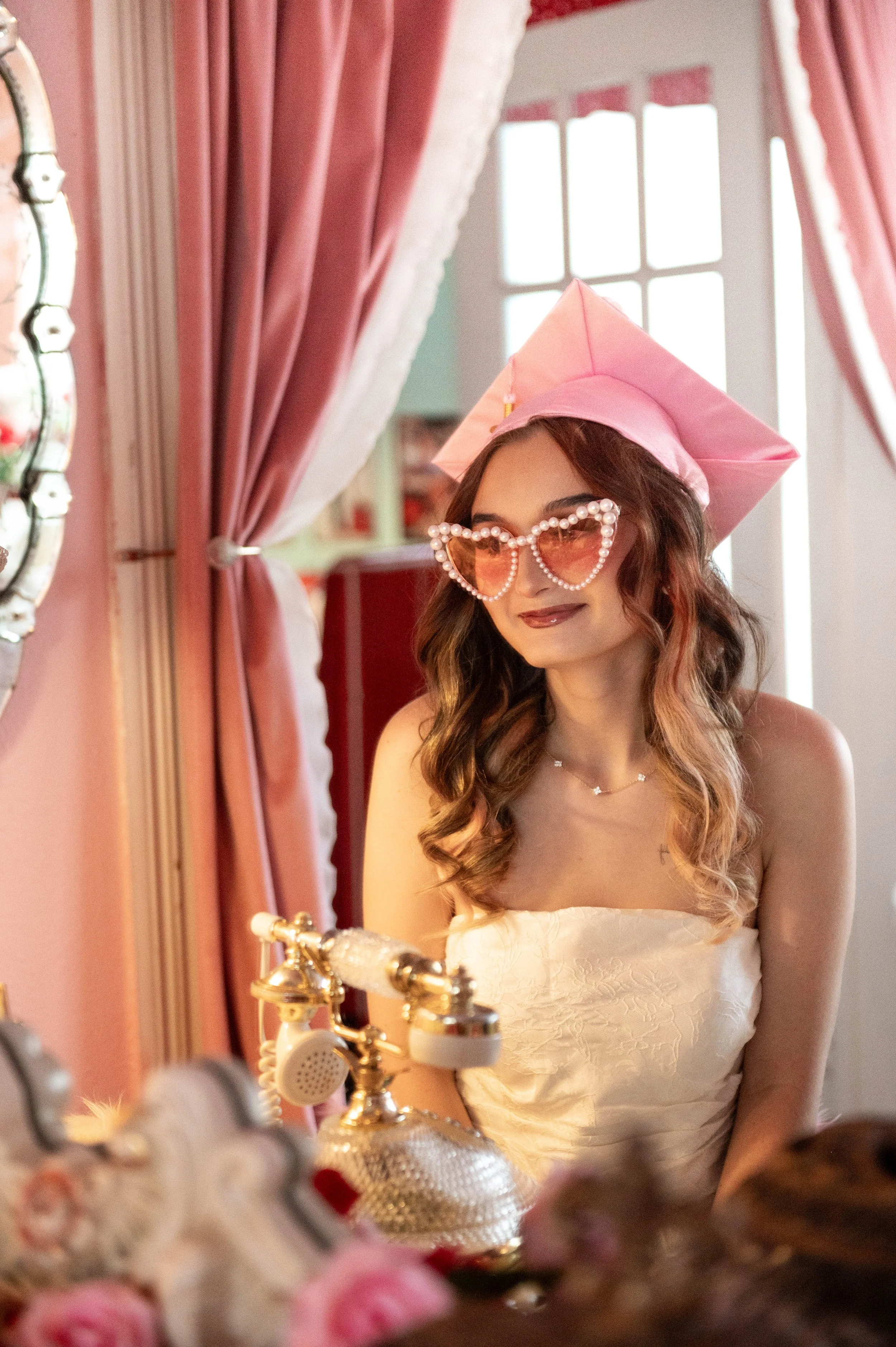 A woman with wavy hair wearing heart-shaped sunglasses with pearl borders, a pink graduation cap, and a white strapless dress looking into a mirror in a room with pink curtains.
