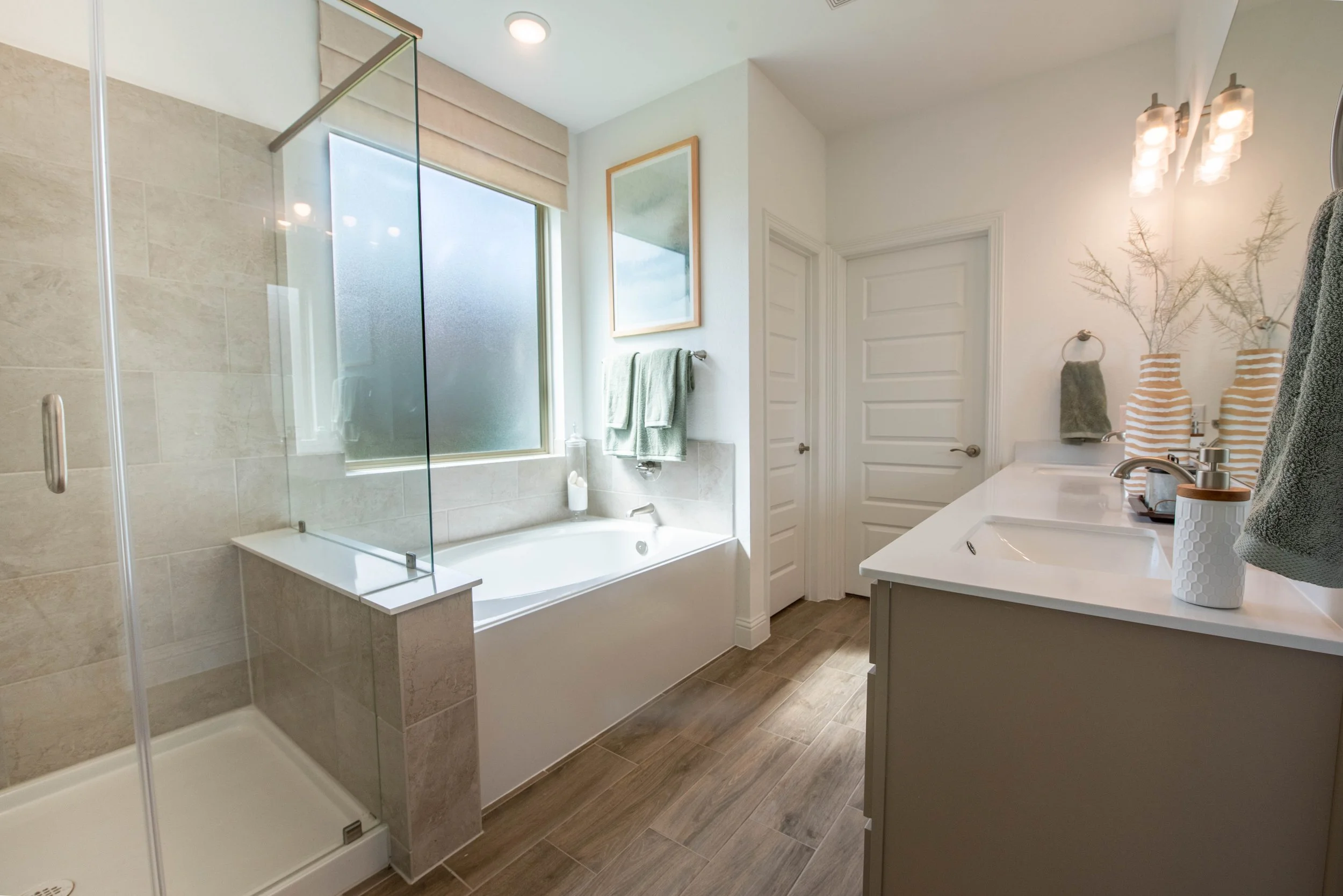Modern bathroom with a walk-in shower with frosted glass, a bathtub next to a large window, and a white vanity with a sink, decorated with striped vases, dried plants, and towels.