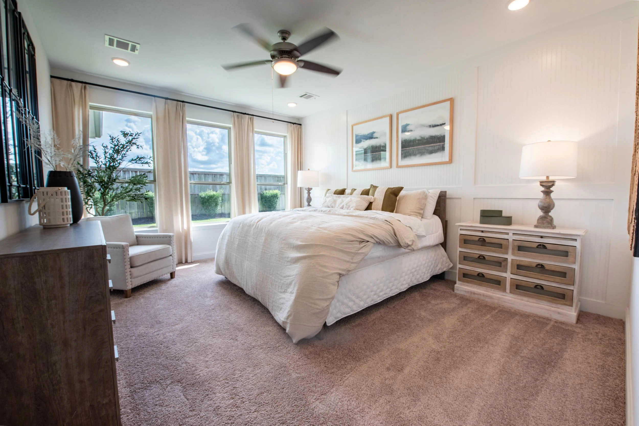 A bedroom with a large bed, nightstands with lamps, framed artwork, a ceiling fan, large windows with curtains, a chair, and a dresser with decorative items.