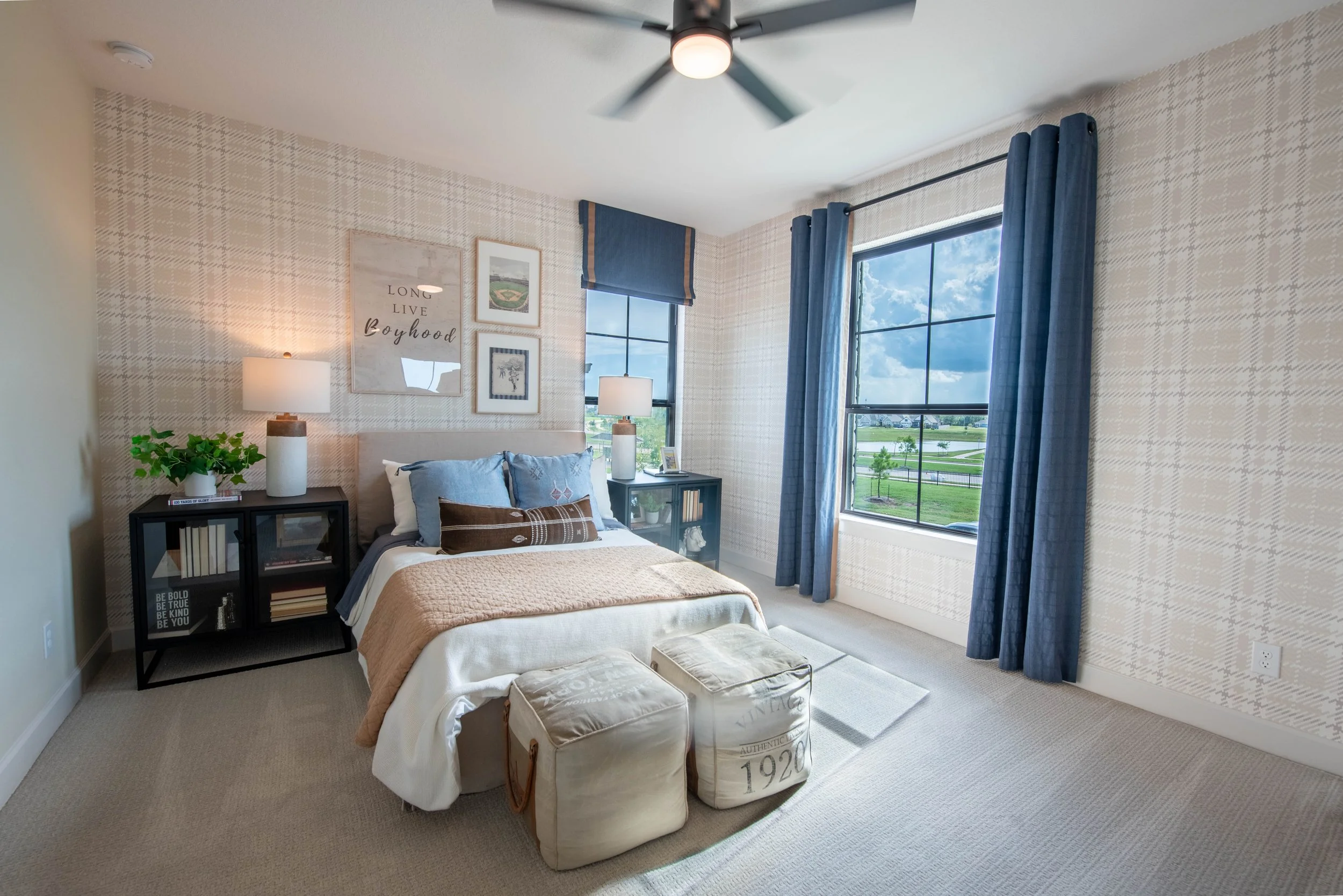 A cozy bedroom with a large window showing a view of a clear blue sky and green landscape. The bed has blue and white bedding, and two matching bedside tables with lamps. The wall behind the bed is decorated with framed art and a small plant. A ceili