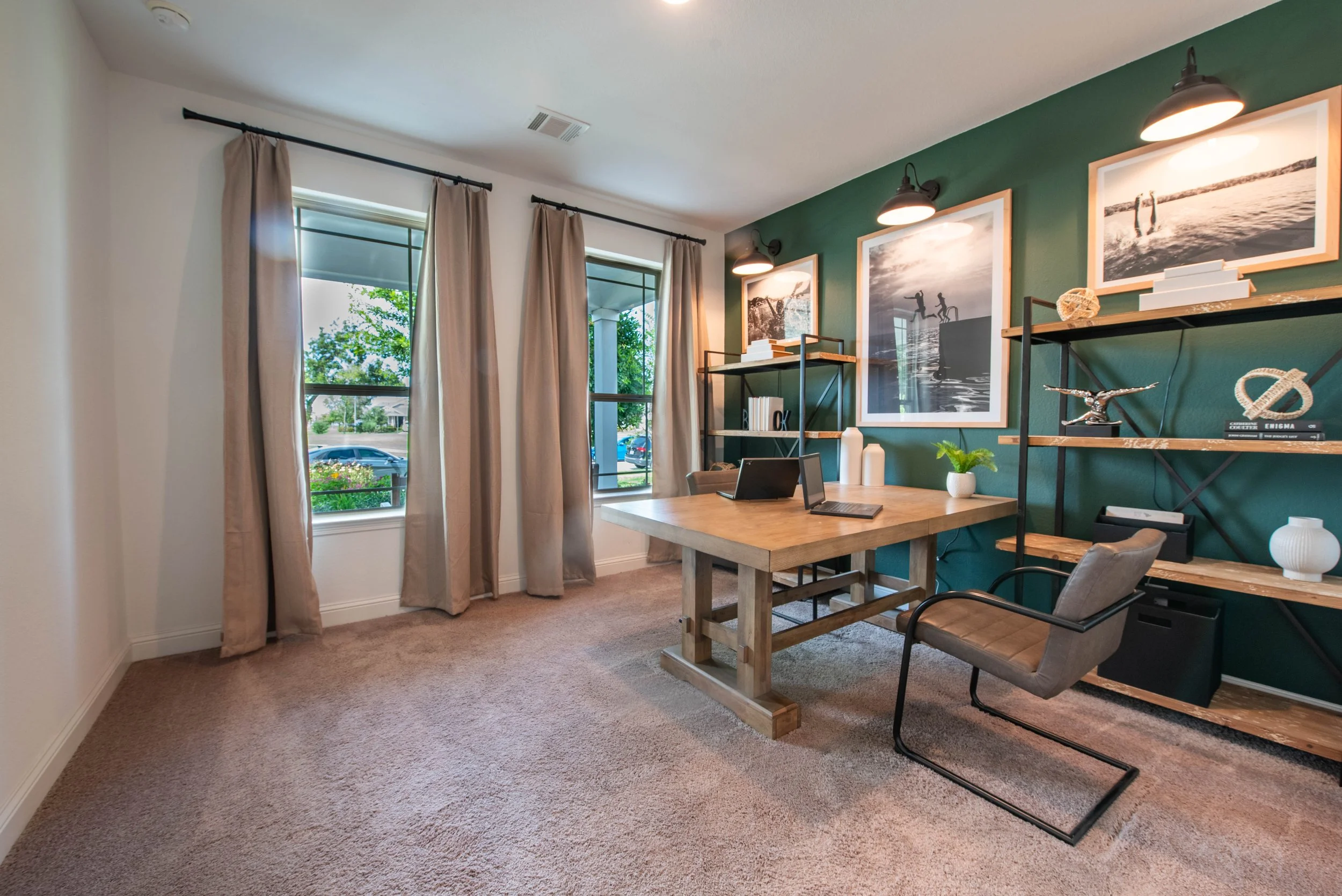 A cozy home office with a green accent wall, framed black and white photographs, a wooden desk, a gray chair, a metal and wood bookshelf with decor, books, and a small potted plant, large windows with beige curtains, and natural light.