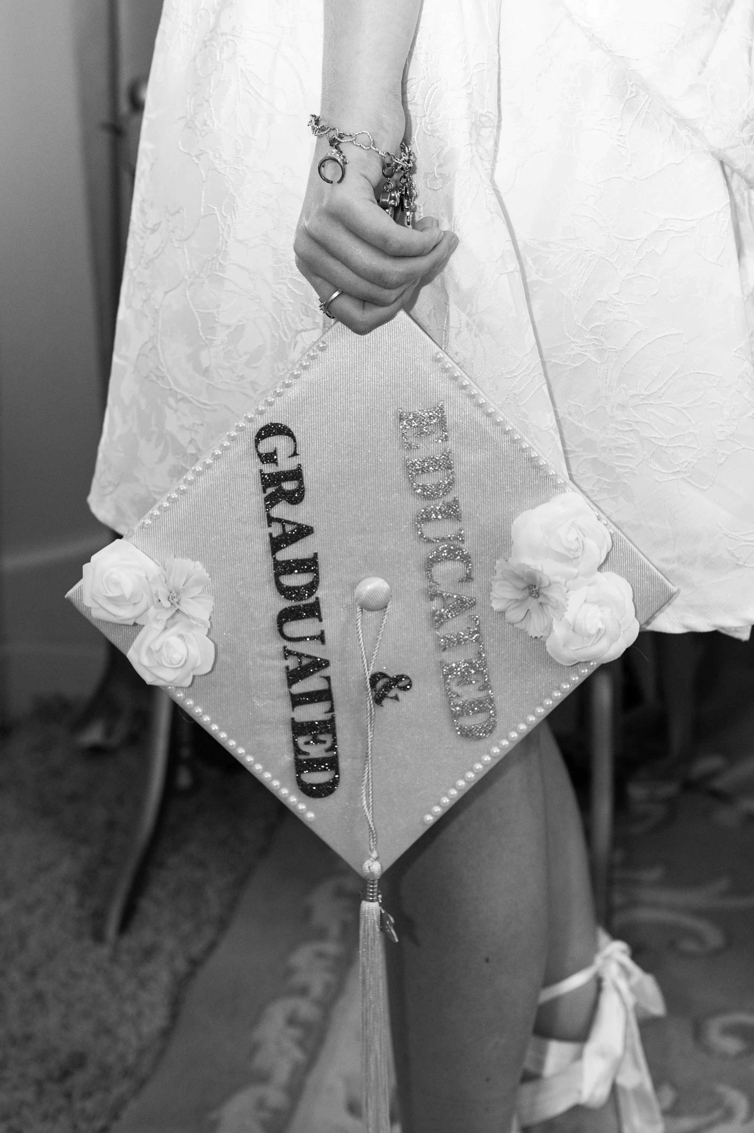 Close-up of a woman's hand holding a decorated pillow with a sash that reads "GROOM'S BEST MAN." The woman is wearing a white dress with a floral textured pattern and has several rings and charms on her wrist. The background shows other dresses and p