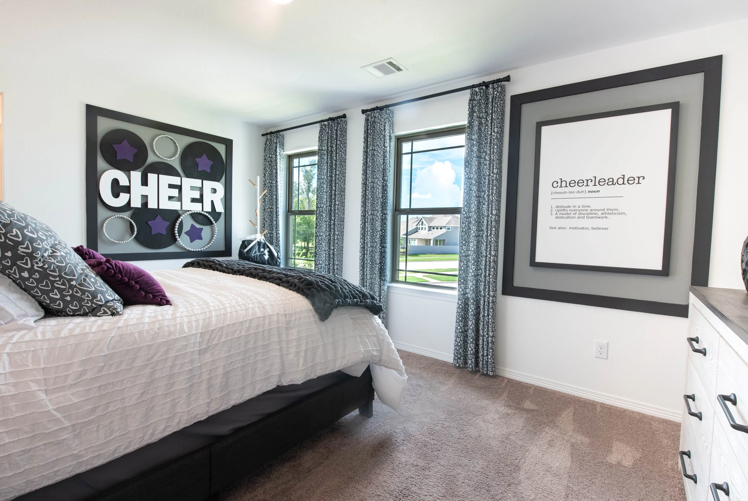 A bedroom with a bed covered in a white textured bedspread, pillows with heart patterns, and a dark blanket. There are two large windows with patterned curtains, and artwork on the wall including a frame with the word 'CHEER' and a definition of 'che