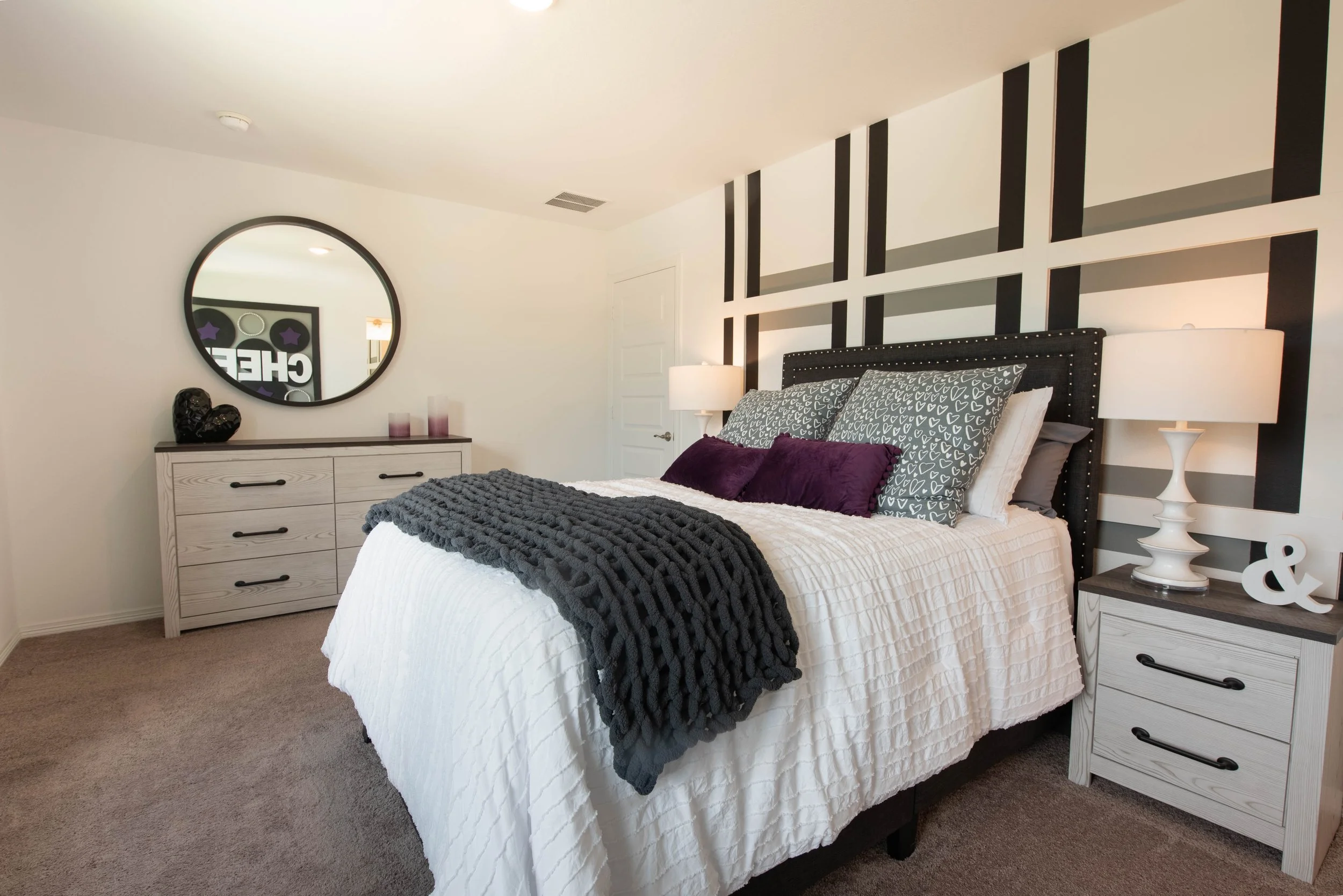 A bedroom with a white bed, black and white geometric pillows, a dark gray throw blanket, and matching white nightstands with lamps. The wall behind the bed has a black and white striped pattern, and there is a mirror and decorative items on a dresse