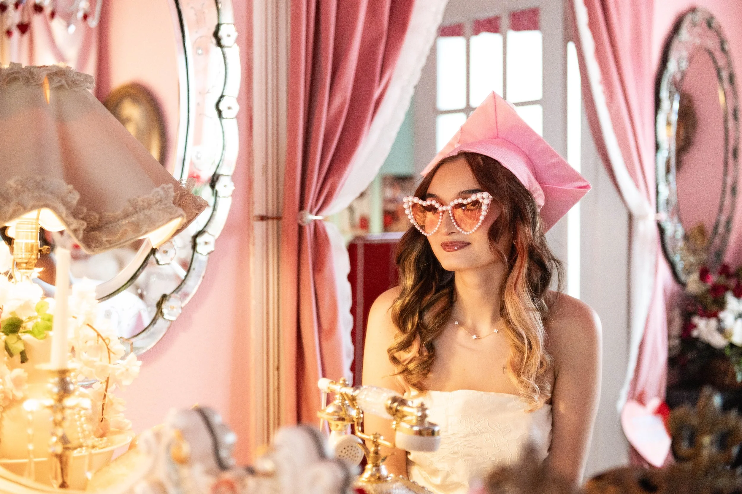 A woman at a vanity table wearing a pink paper crown and heart-shaped pearl sunglasses, in a room with pink curtains and vintage mirrors.
