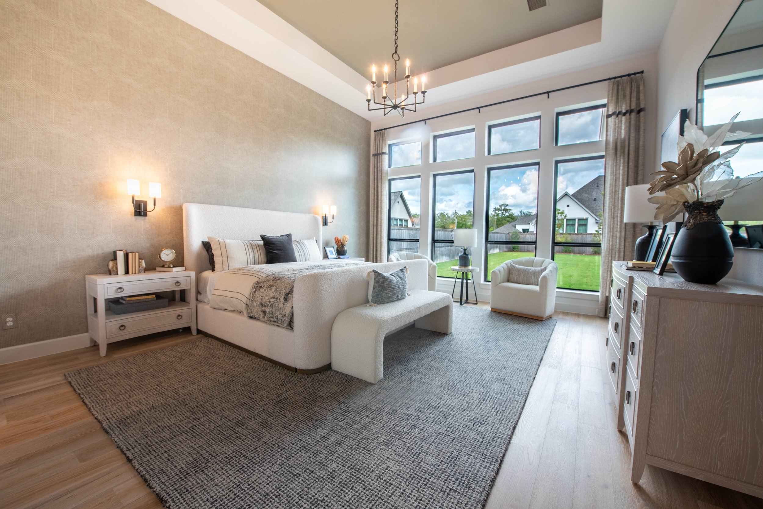 A spacious modern bedroom with large windows showing a backyard and houses outside. It has a white bed with multiple pillows, a white side table with books, a clock, and decor, and contains a black vase with decorative flowers on a white dresser. The