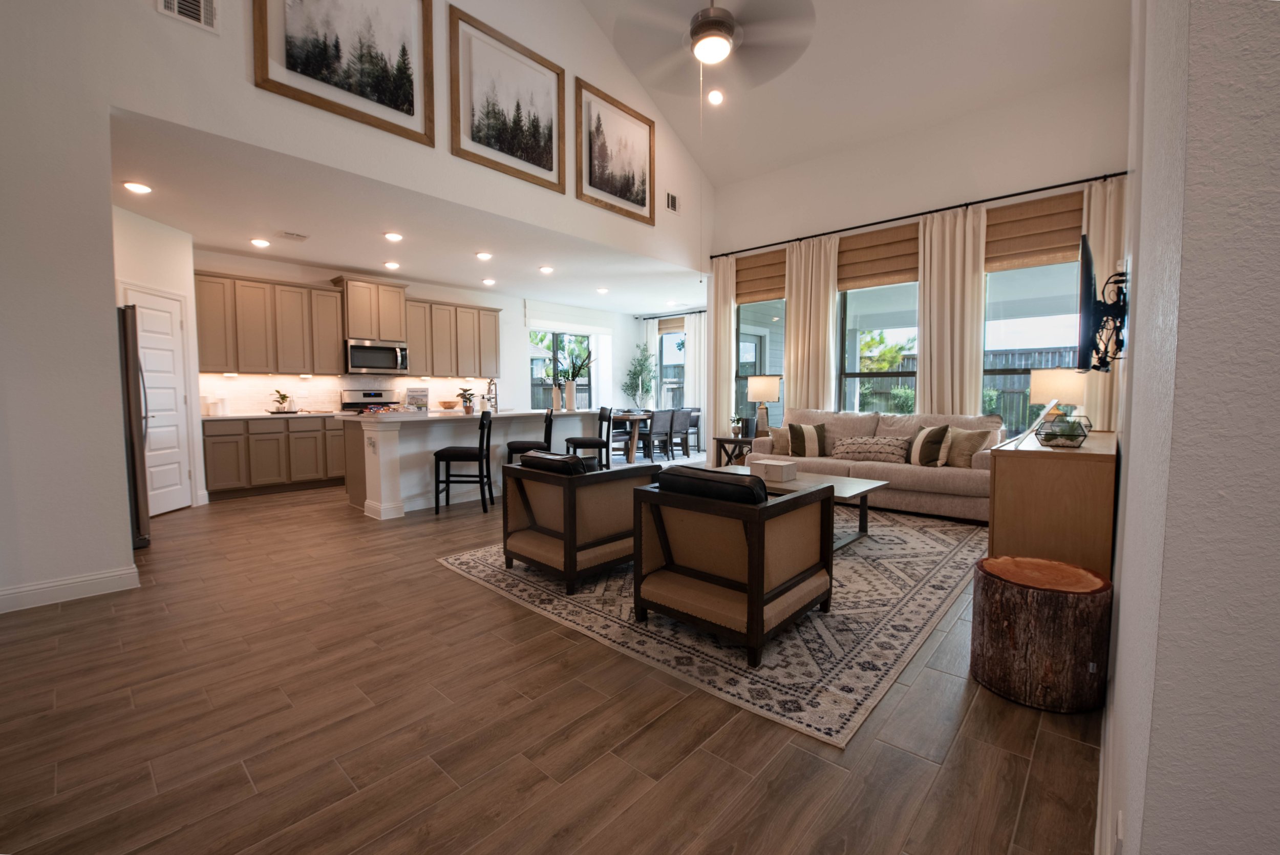 Open-concept living room and kitchen area with large windows, beige sofa, armchairs, patterned rug, and wooden accents.
