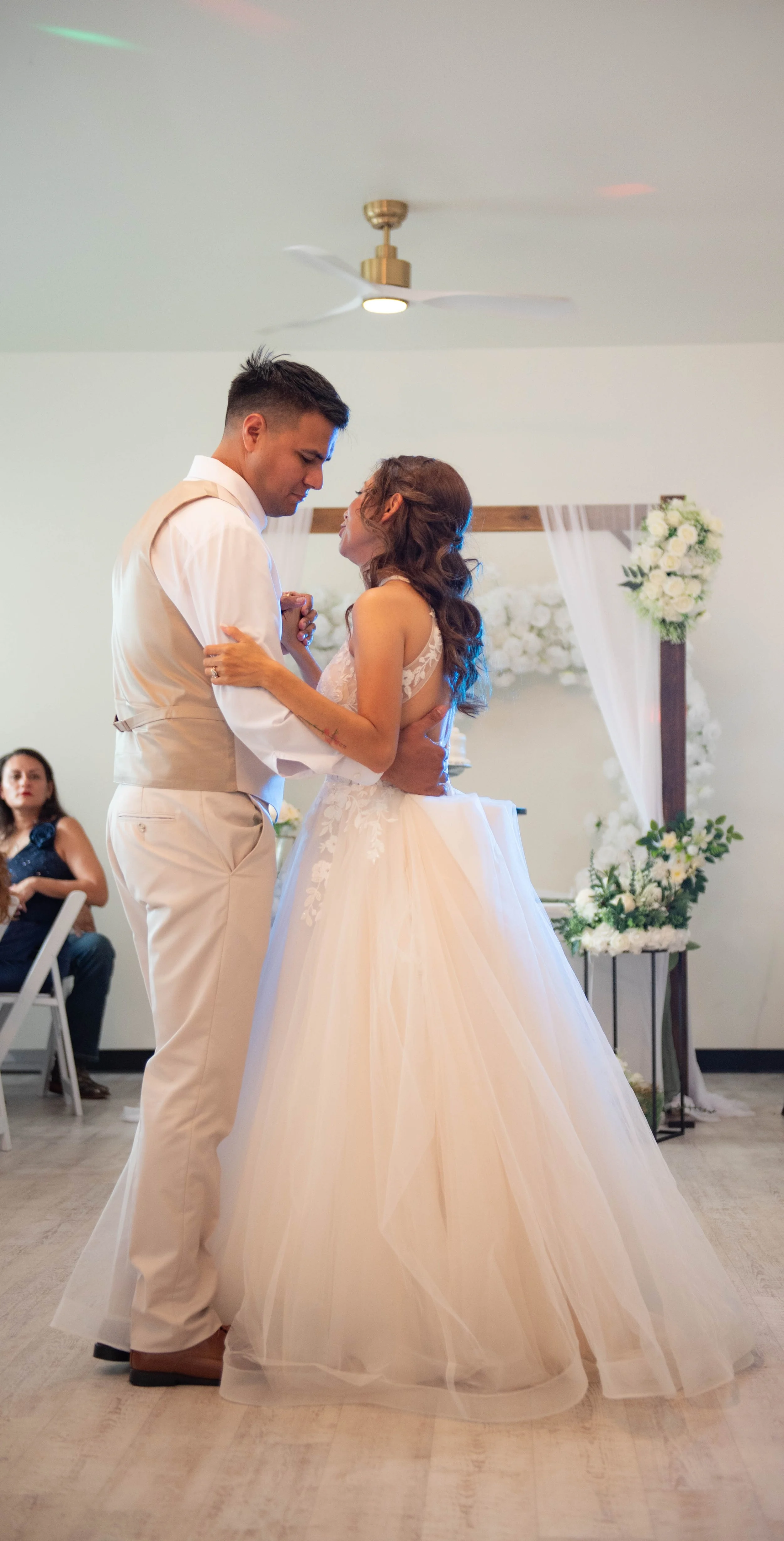 A bride and groom are dancing during their wedding reception in a decorated room with white flowers and a wooden arch in the background.