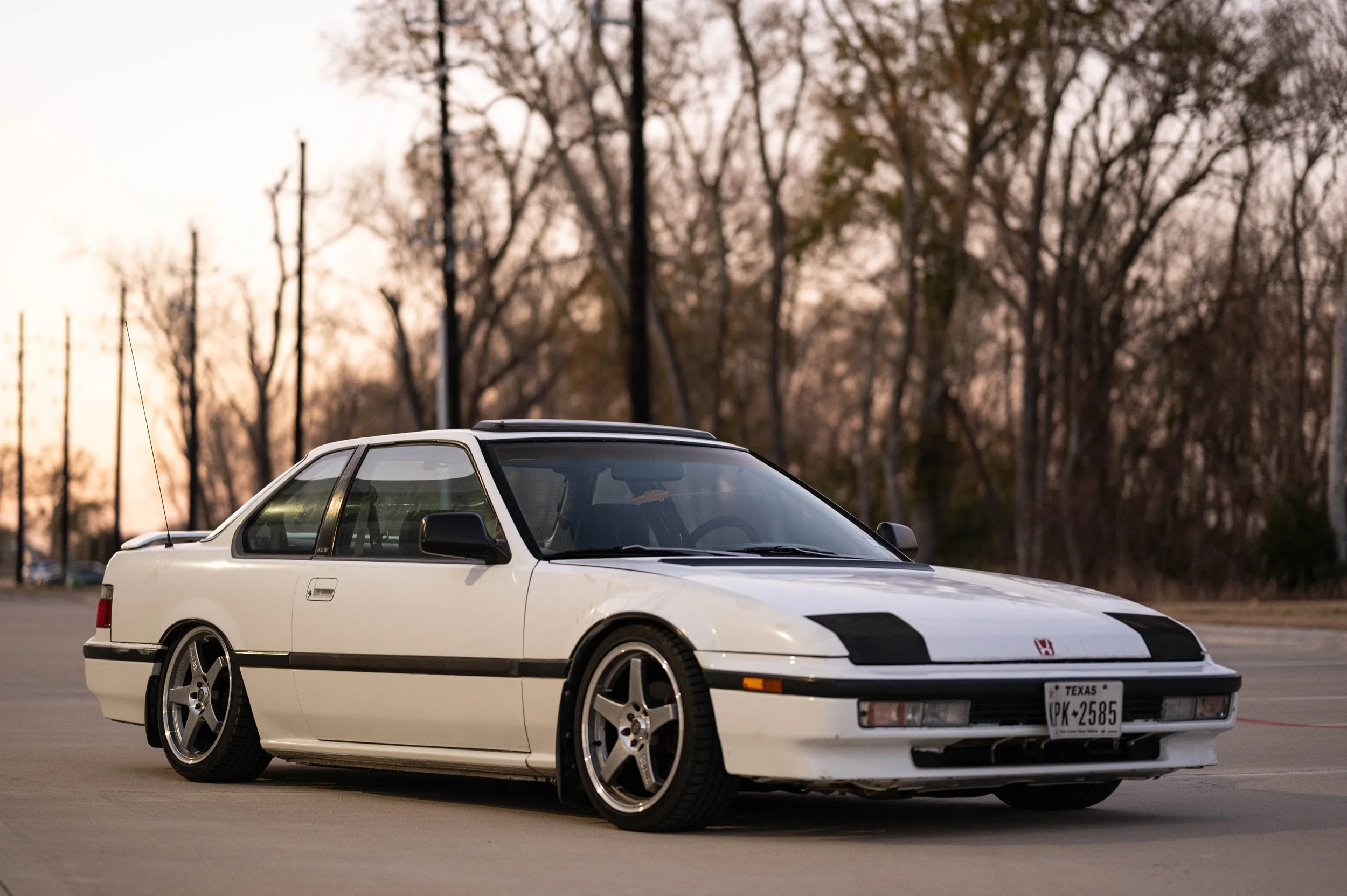 A white sports car with a black stripe on the hood, parked in an open lot during sunset with bare trees in the background.