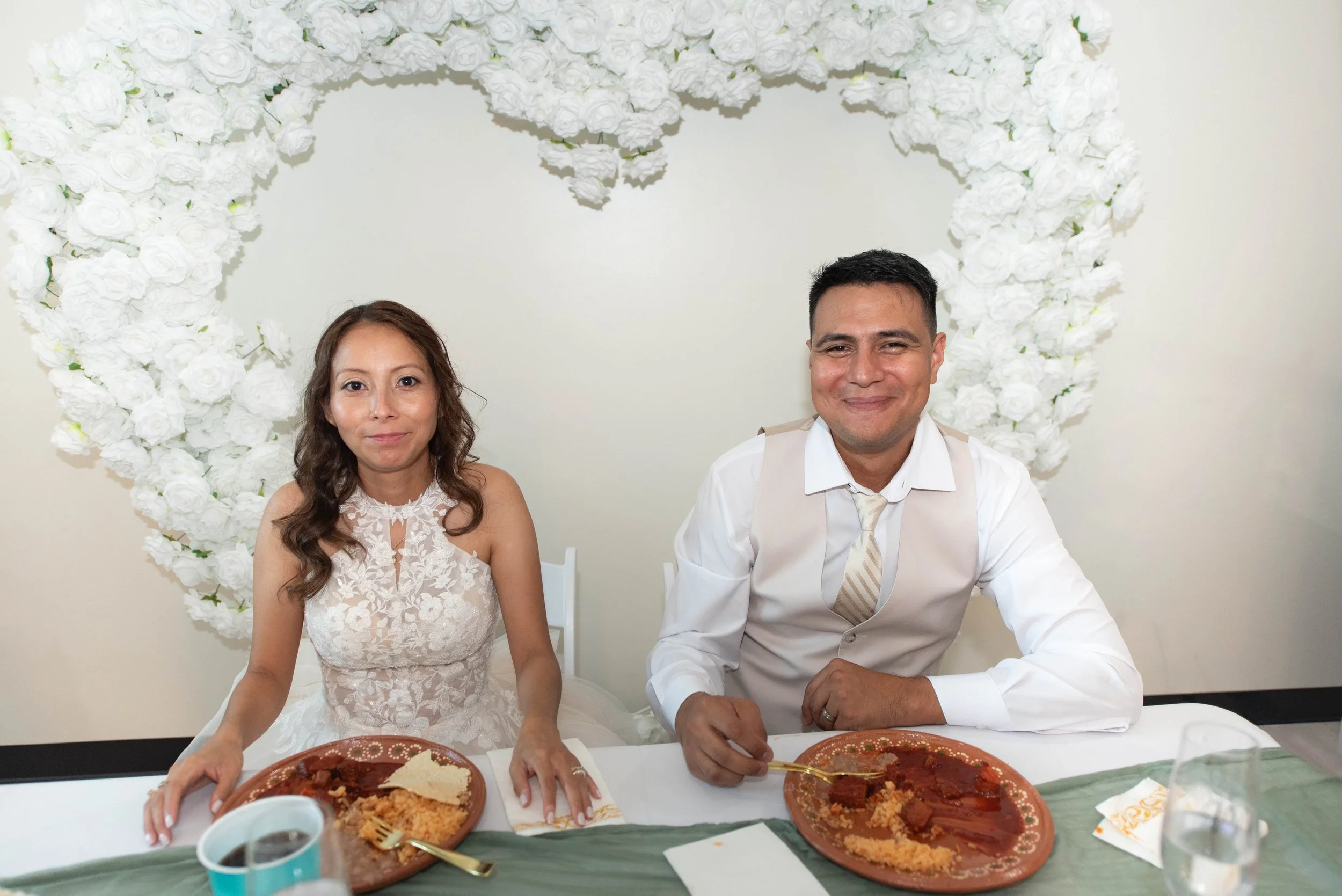 A couple sitting at a table during a wedding reception, with a heart-shaped floral arrangement in the background, eating food from plates.