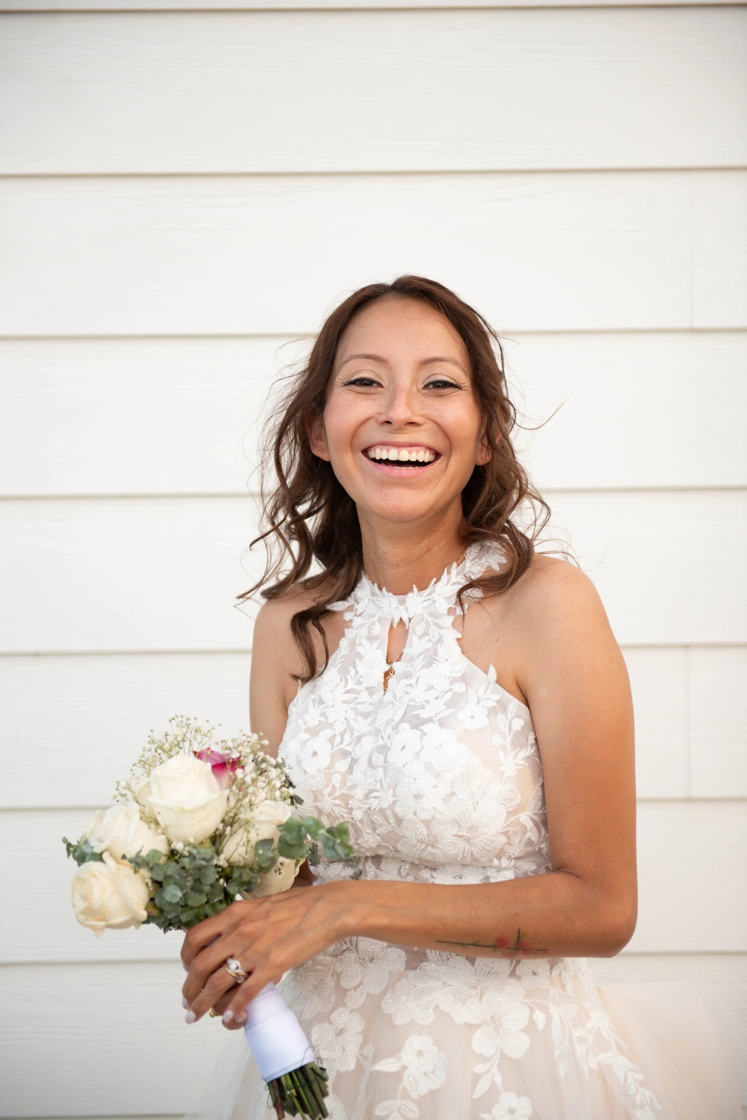 A smiling woman with brown hair in a wedding dress holding a bouquet of white and pink roses and greenery, standing in front of a white wooden wall.