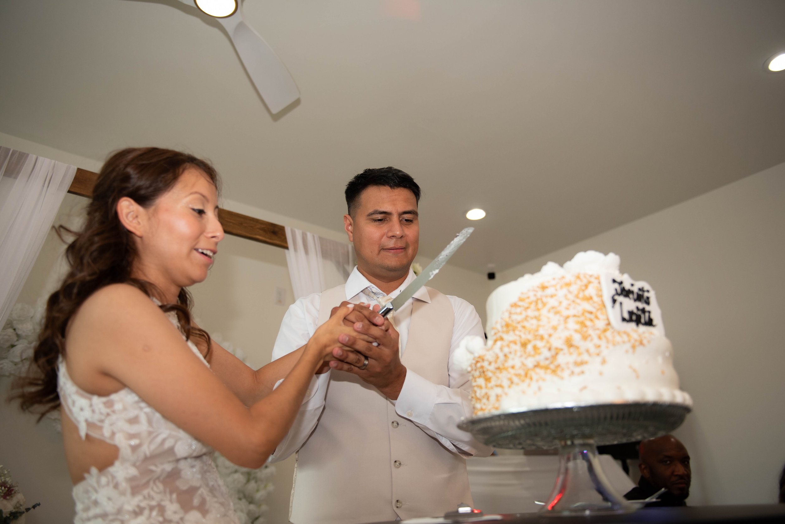 A newlywed couple cutting their wedding cake together at their reception, with a wedding cake decorated with white frosting and orange sprinkles in the foreground.