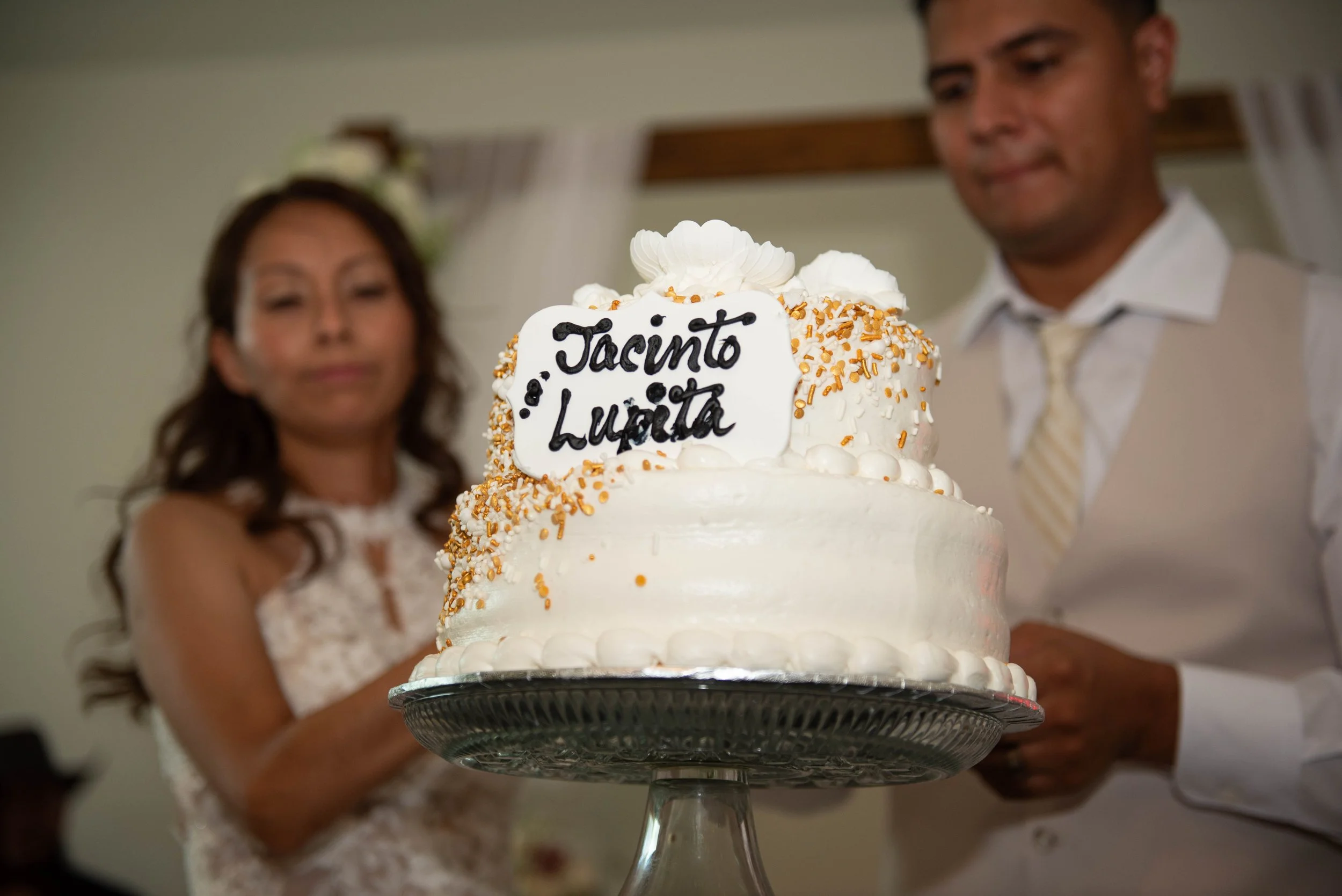 A wedding cake with white frosting, gold sprinkles, and a white chocolate plaque with the message 'Tacinto & Luzta' written in black. The cake is on a silver stand, and a bride and groom are blurred in the background.