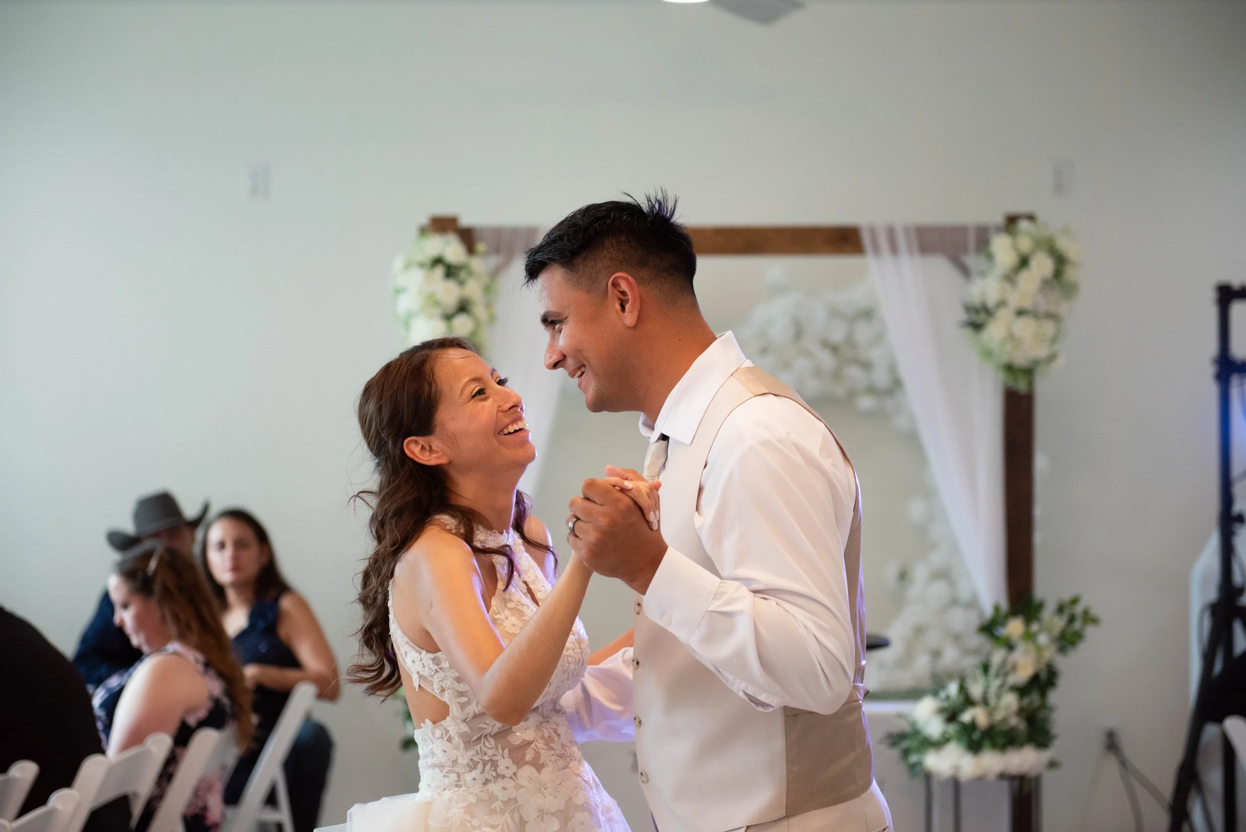 A bride and groom dance closely together during their wedding reception, with the bride smiling and looking up at the groom in front of a decorated backdrop.
