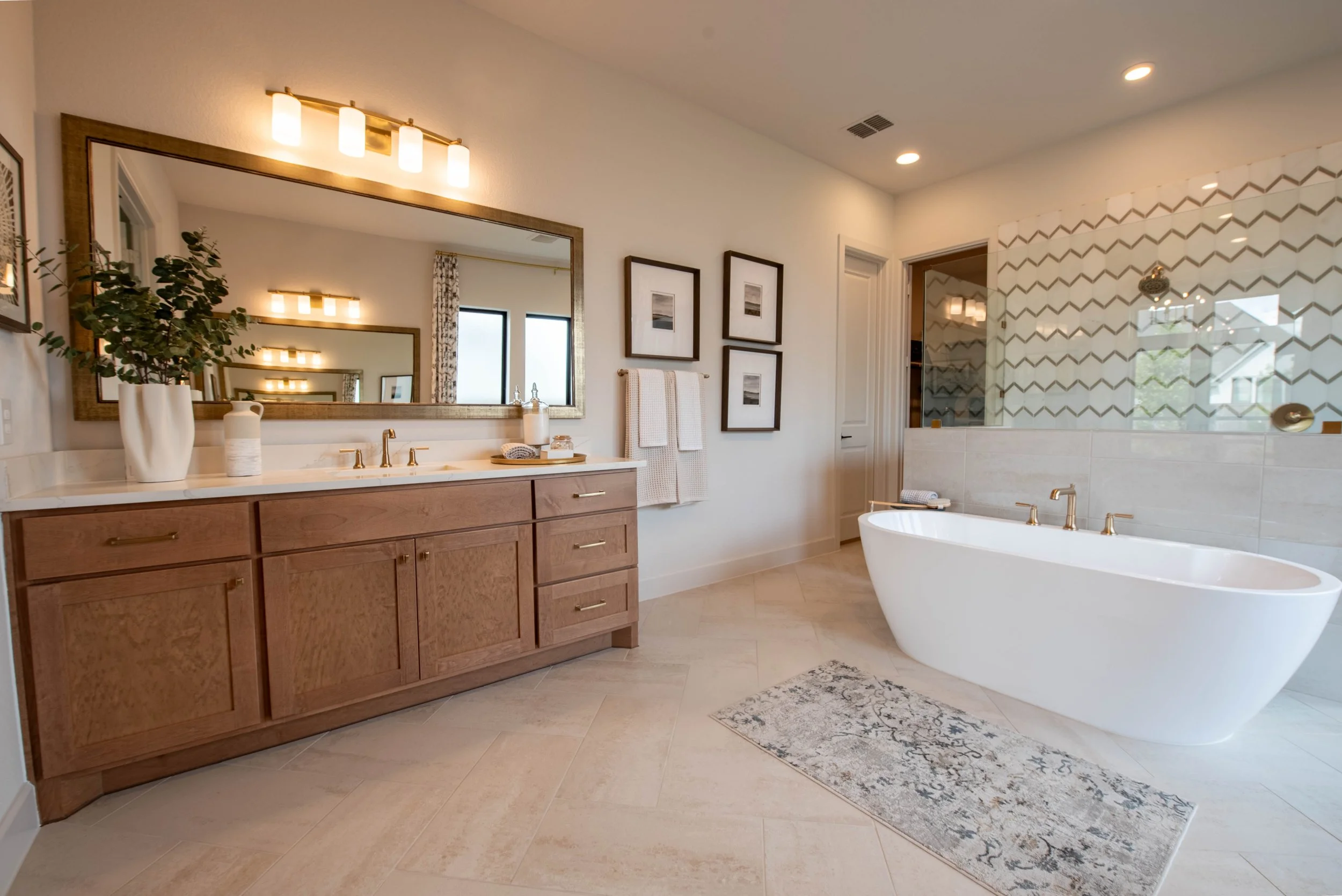 Modern bathroom with a large mirror above a wooden vanity, a bathtub with gold fixtures, and a walk-in shower with patterned glass, beige floor tiles, and framed wall art.