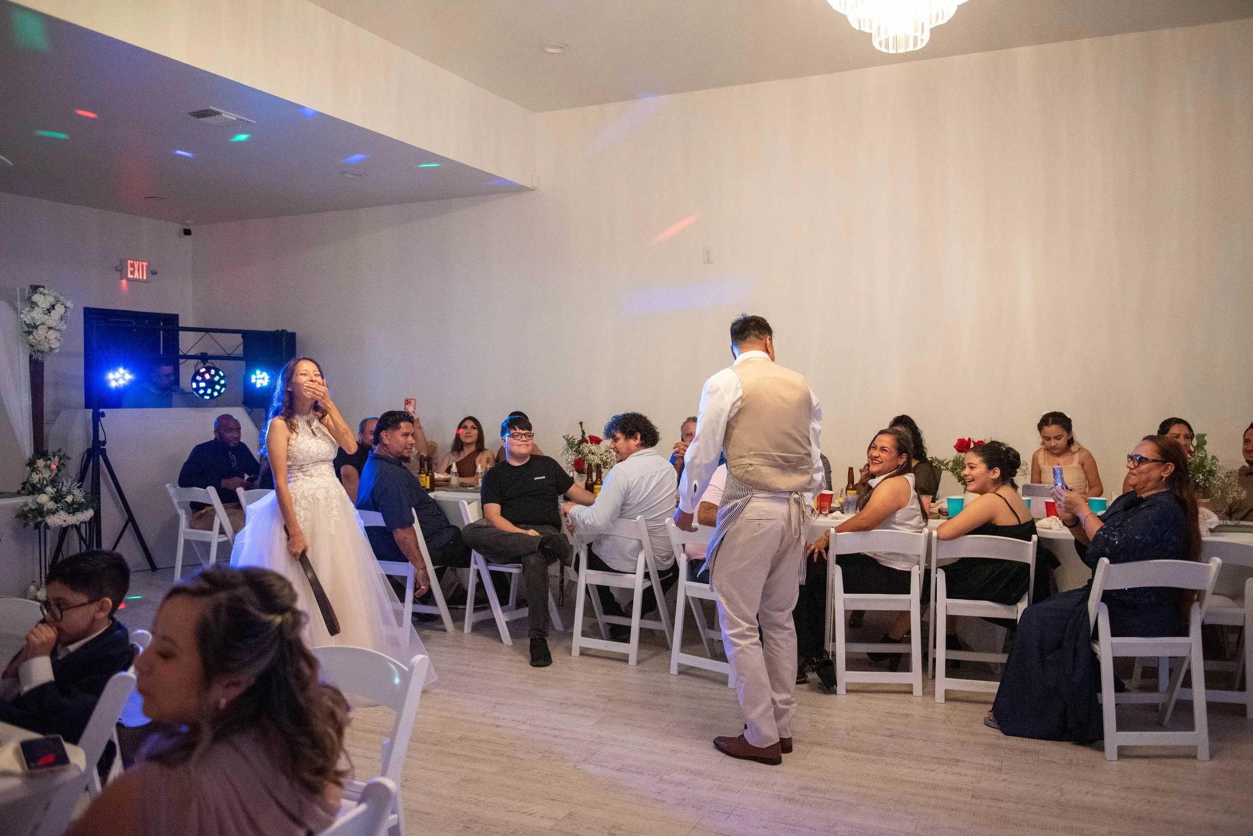 A wedding reception scene with a bride in a white wedding dress and veil, laughing, surrounded by guests seated at tables. A man in a beige vest and white shirt holds a microphone, possibly giving a speech. The room is decorated with flowers, and the