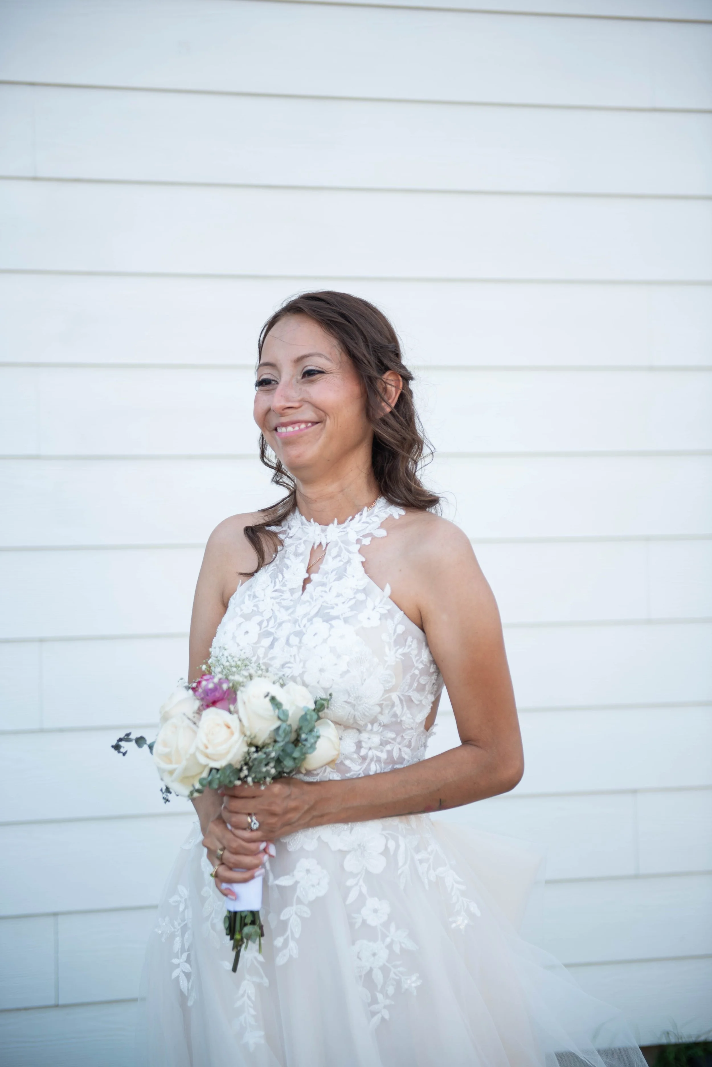 A woman in a white wedding dress holding a bouquet of white and pink flowers, standing in front of a white wooden wall, smiling.