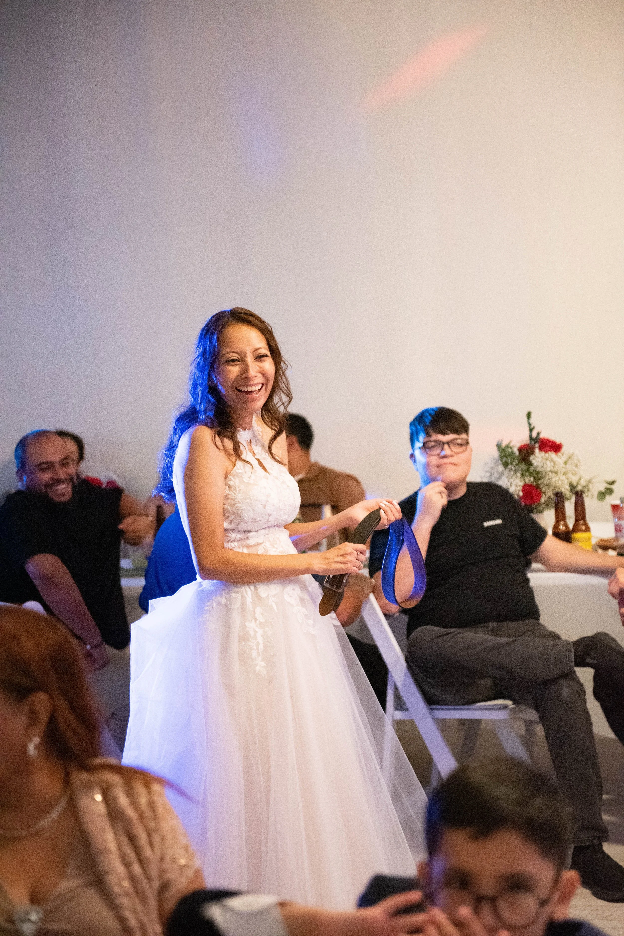 A woman in a white wedding dress smiling and holding a bone-shaped award, seated at a table with other guests in a celebration setting.
