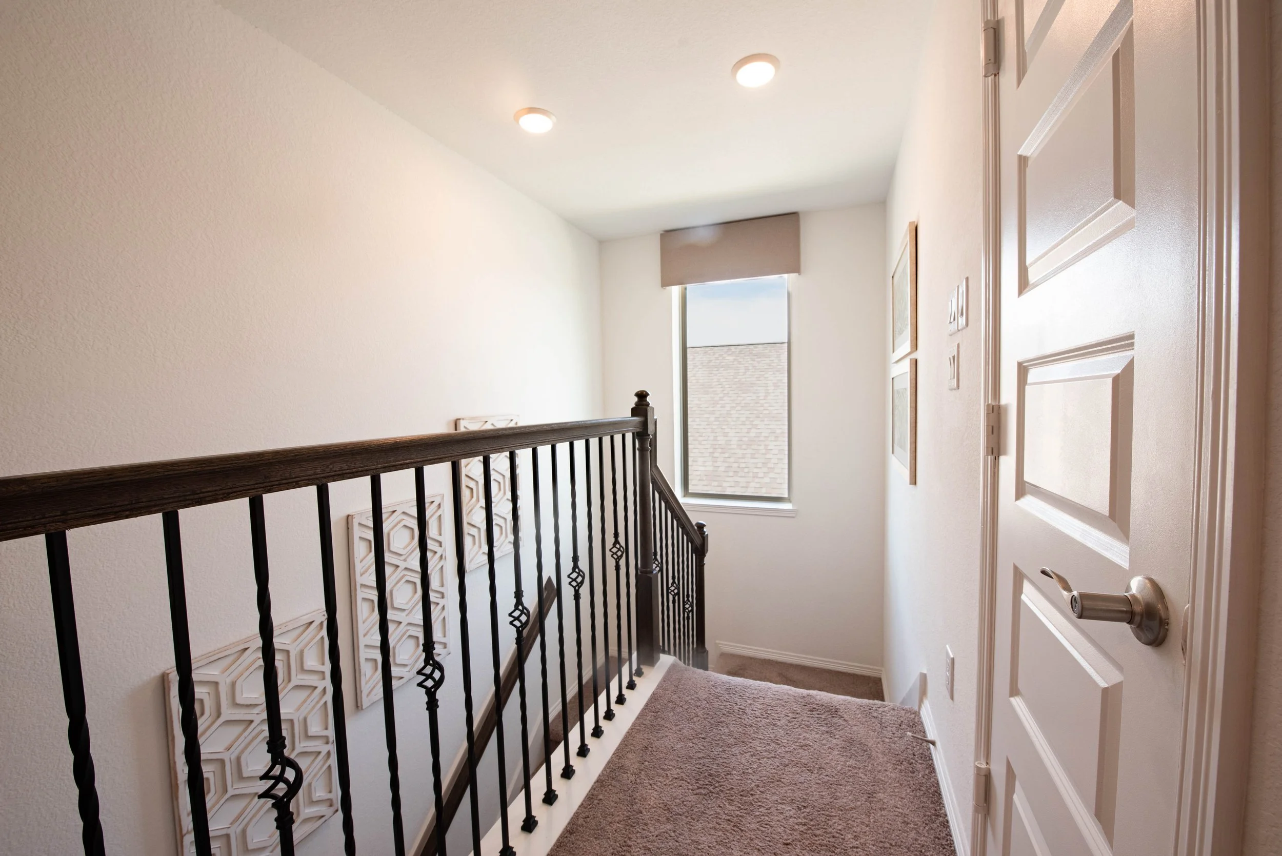 An upstairs hallway with beige walls, carpeted floor, a white door, and a window with a valance letting in natural light. A decorative wall art is on the wall, and a black metal railing runs along the staircase.
