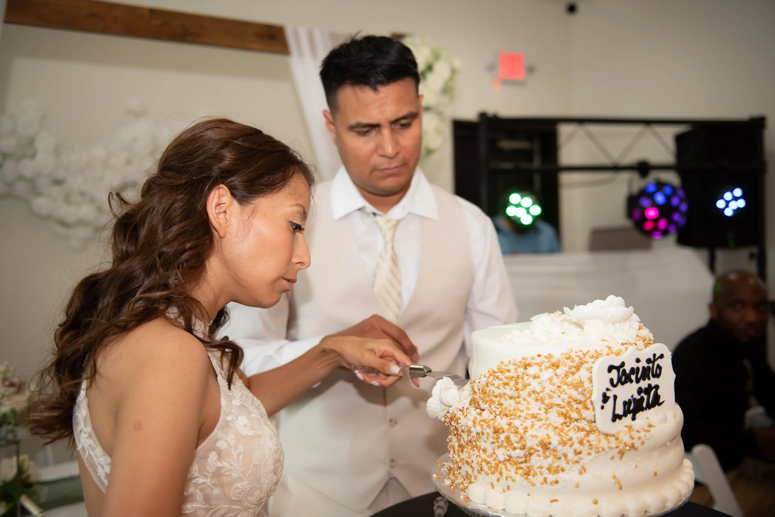 A woman with brown, curly hair in a white lace dress is cutting a wedding cake. A man in a white shirt and beige vest stands next to her, looking at the cake. The cake is decorated with white frosting and orange sprinkles, with a white plaque that ha