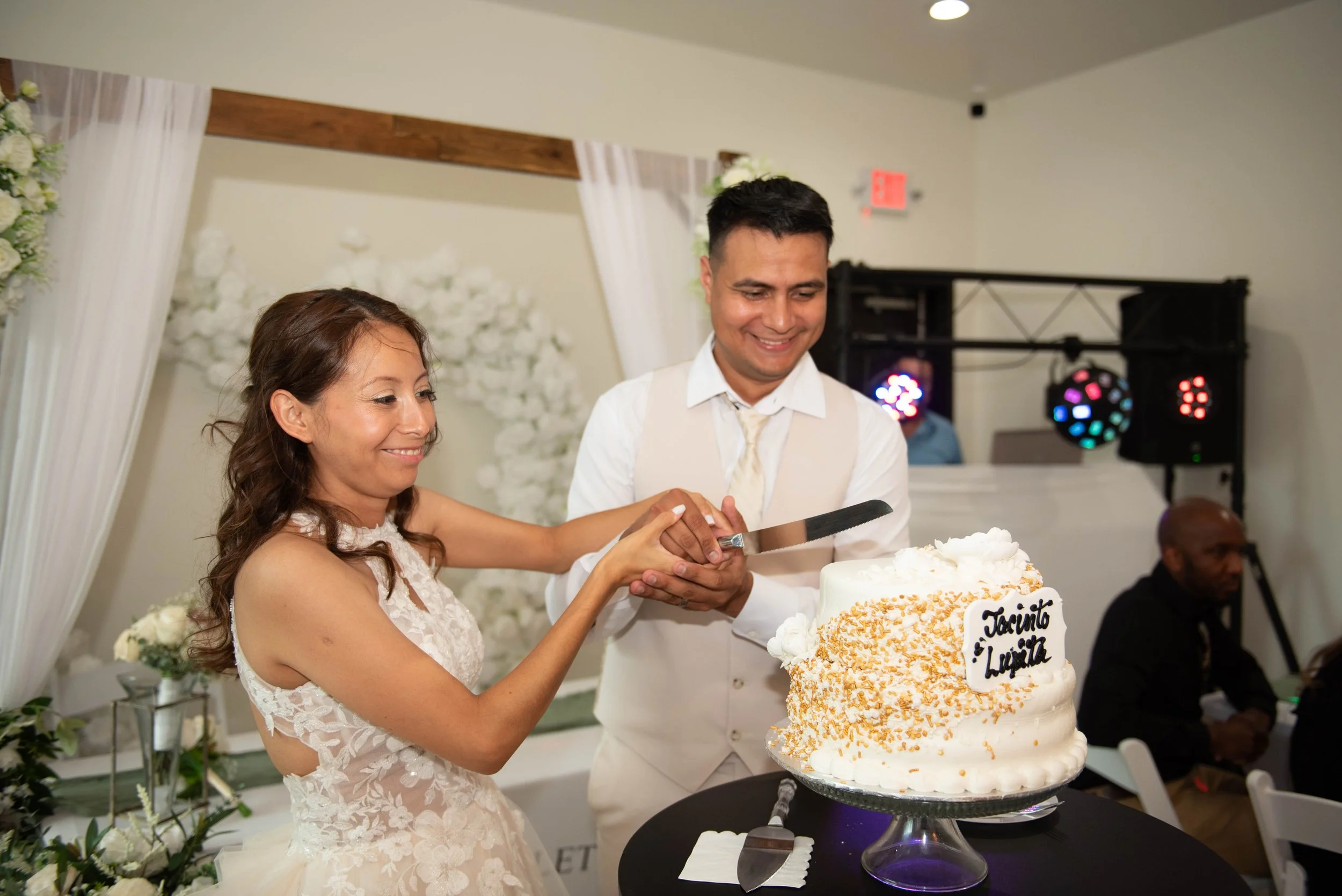 A bride and groom cut a wedding cake together at a reception, with a floral backdrop and a DJ booth in the background.