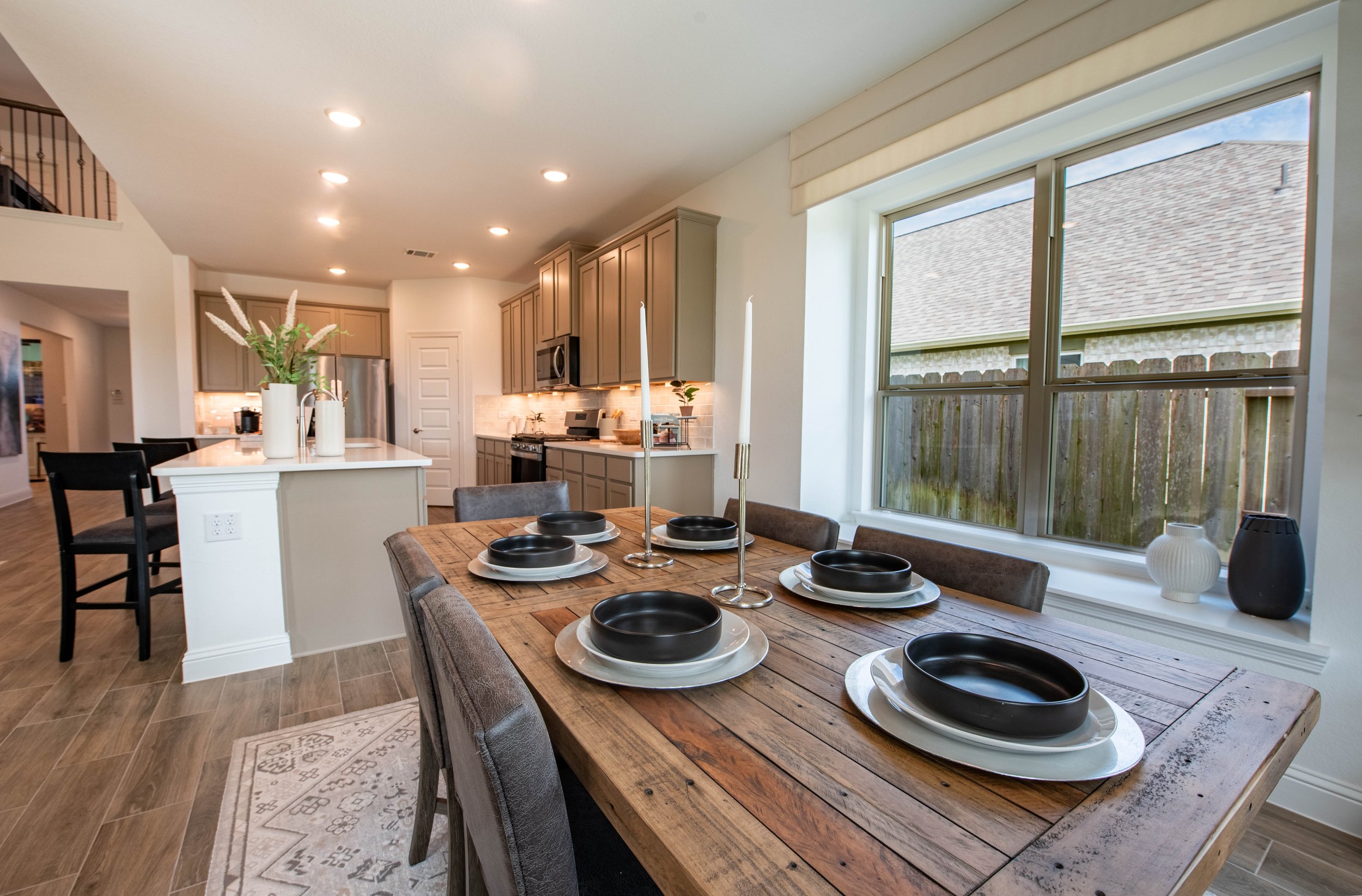 Bright kitchen and dining area with a wooden table set with black bowls and white plates; large window showing a wooden fence and neighboring house; beige cabinets and modern appliances.