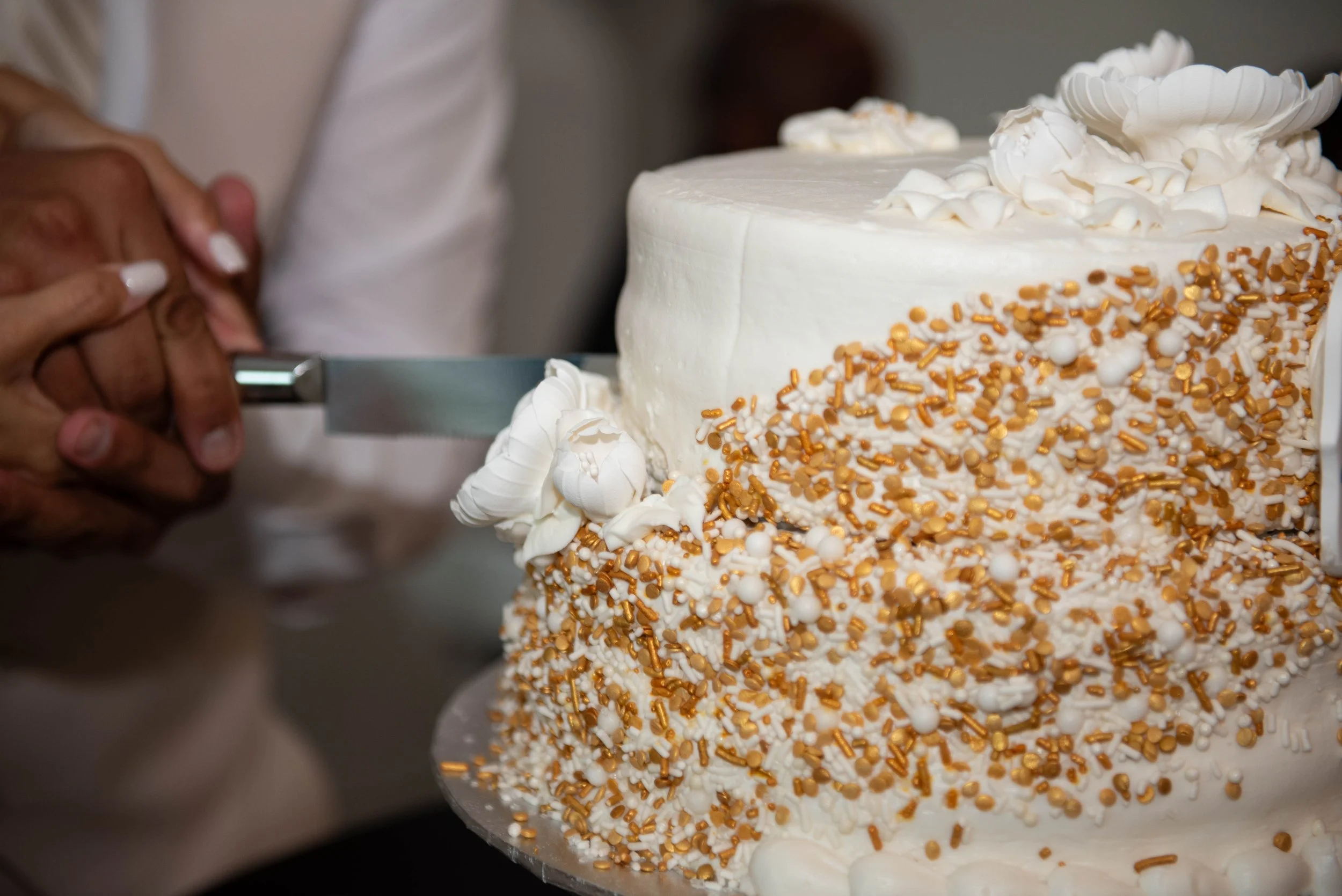 A person decorates a white cake with gold and white sprinkles and icing flowers.