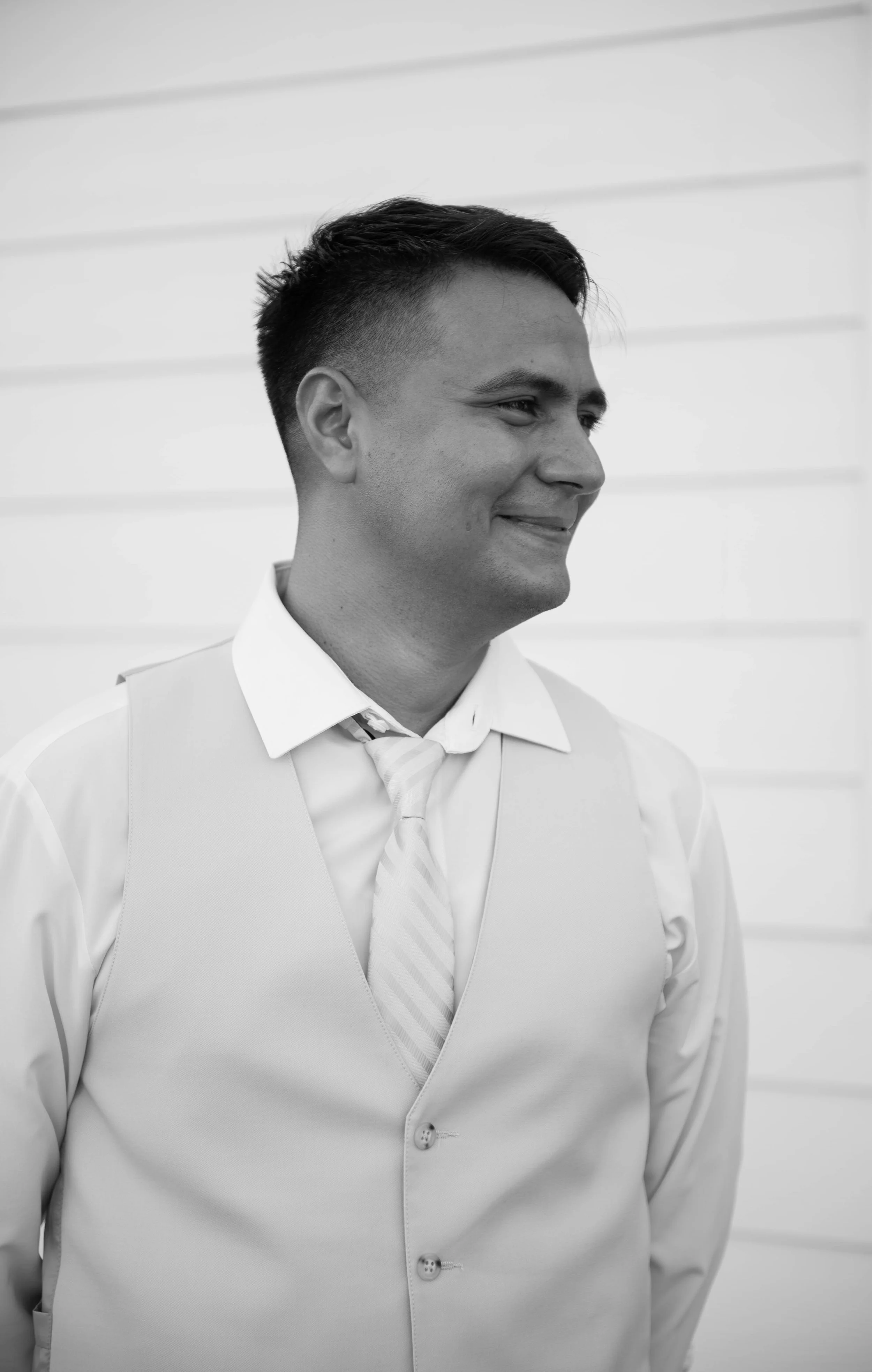 Black and white photo of a man smiling, dressed in a light-colored vest, shirt, and tie, standing against a background of horizontal siding.