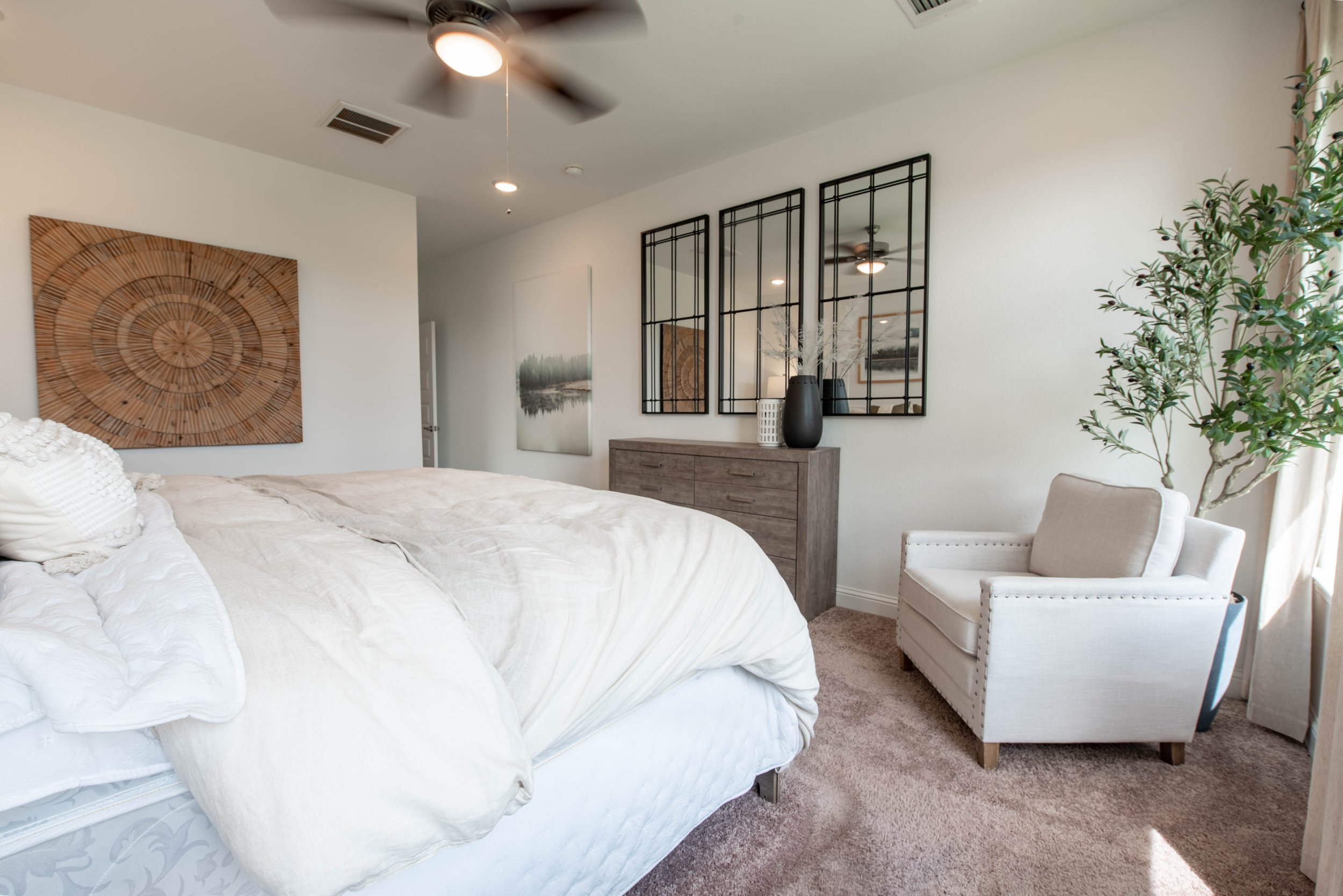 Bedroom with white bed, wooden dresser, large wall art, three decorative mirrors, plants, and a chair.