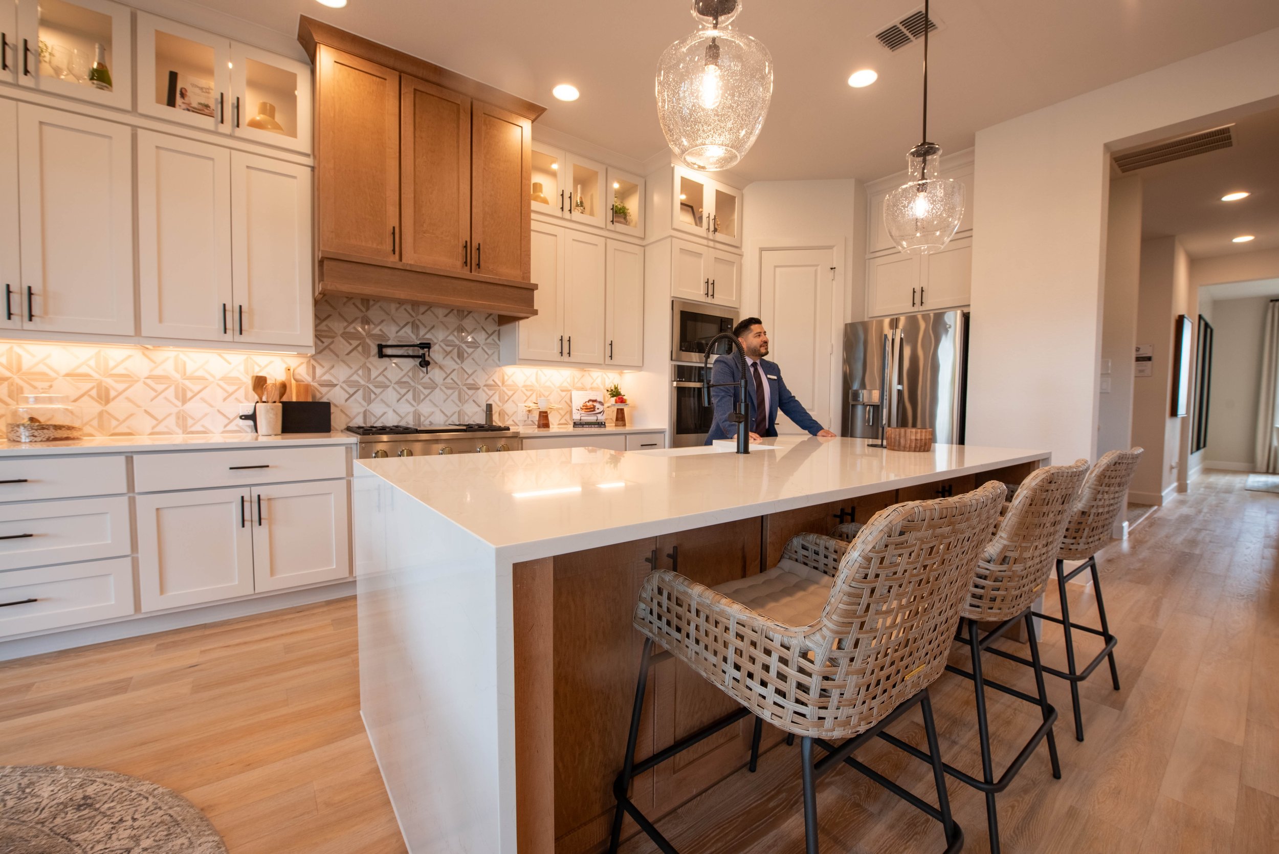 Modern kitchen with white and wood cabinetry, a large island with a white countertop, wicker bar stools, and two pendant lights hanging from the ceiling. A man in a suit is standing at the kitchen island.
