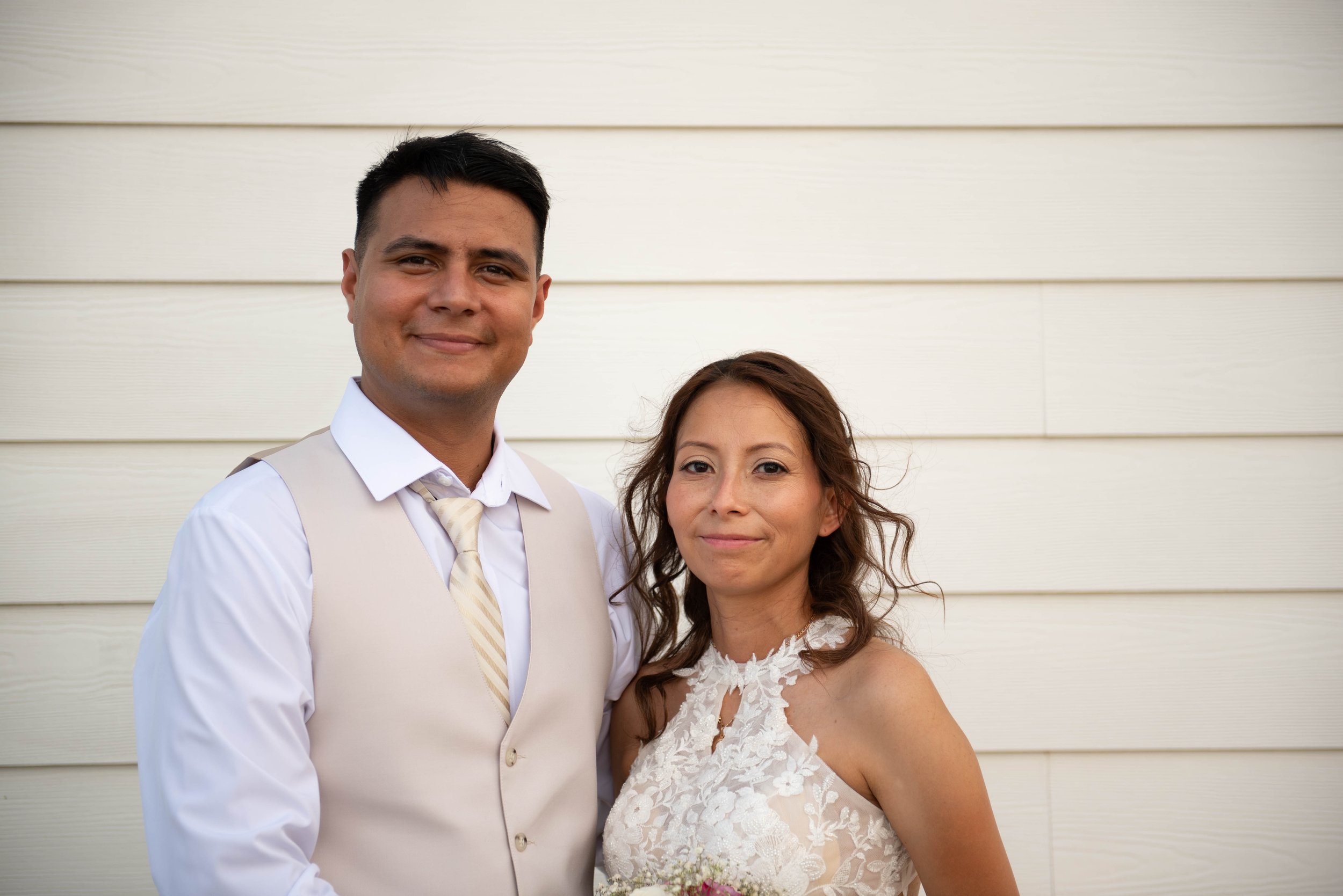 A man and a woman pose together outside against a white horizontal siding wall. The man is wearing a white shirt, beige vest, and a striped tie, and the woman is dressed in a white lace dress holding a bouquet.
